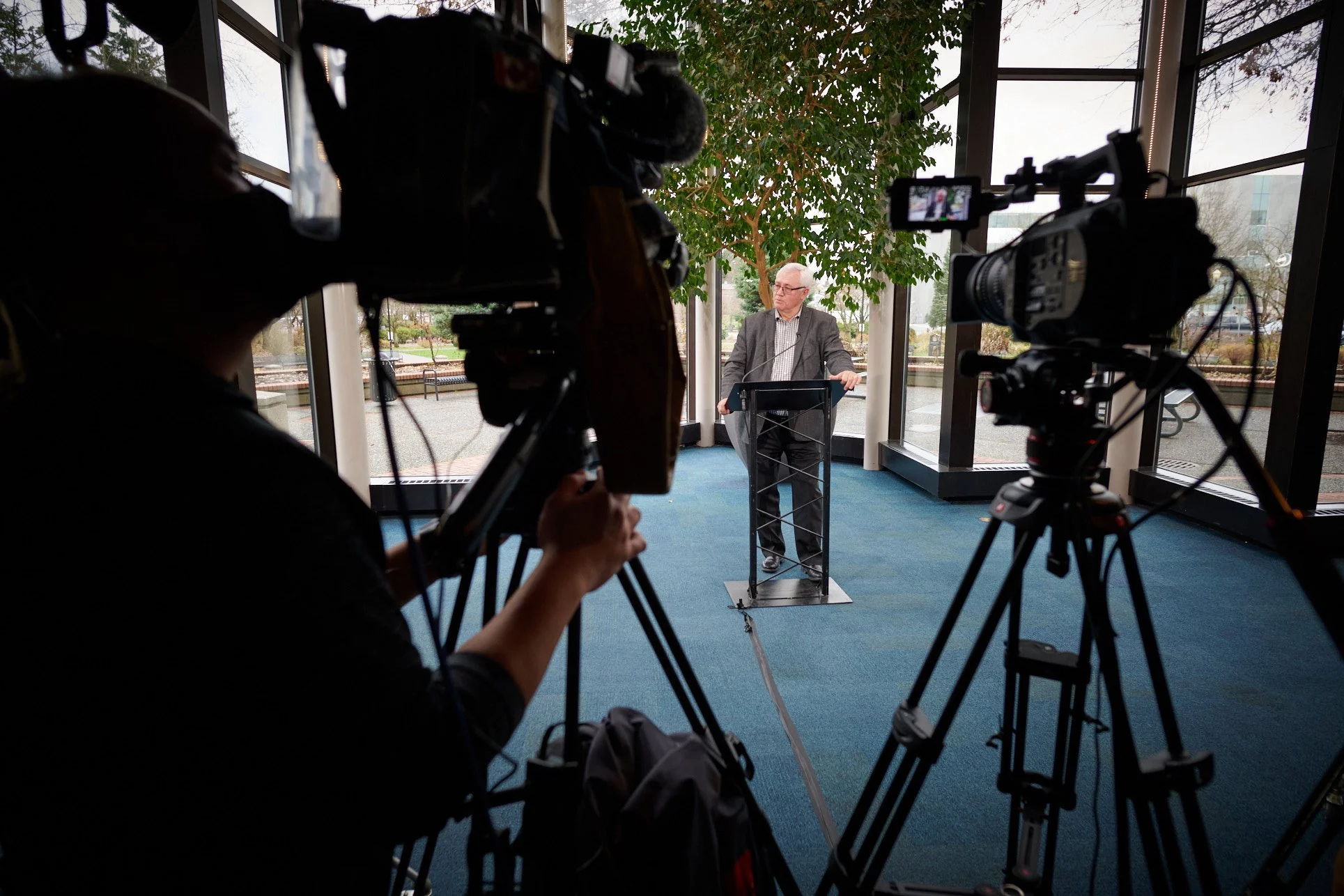 A man in a suit and glasses stands at a podium giving a speech or presentation inside a building with large windows and trees outside. There are cameras recording him, with a silhouette of a cameraman in the foreground.