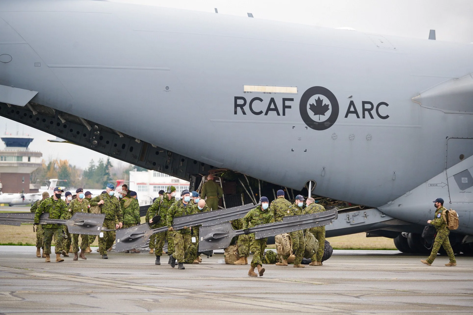 Military personnel in camouflage uniforms and face masks walk alongside a gray military aircraft with the markings RCAF ARC, on an airport tarmac.