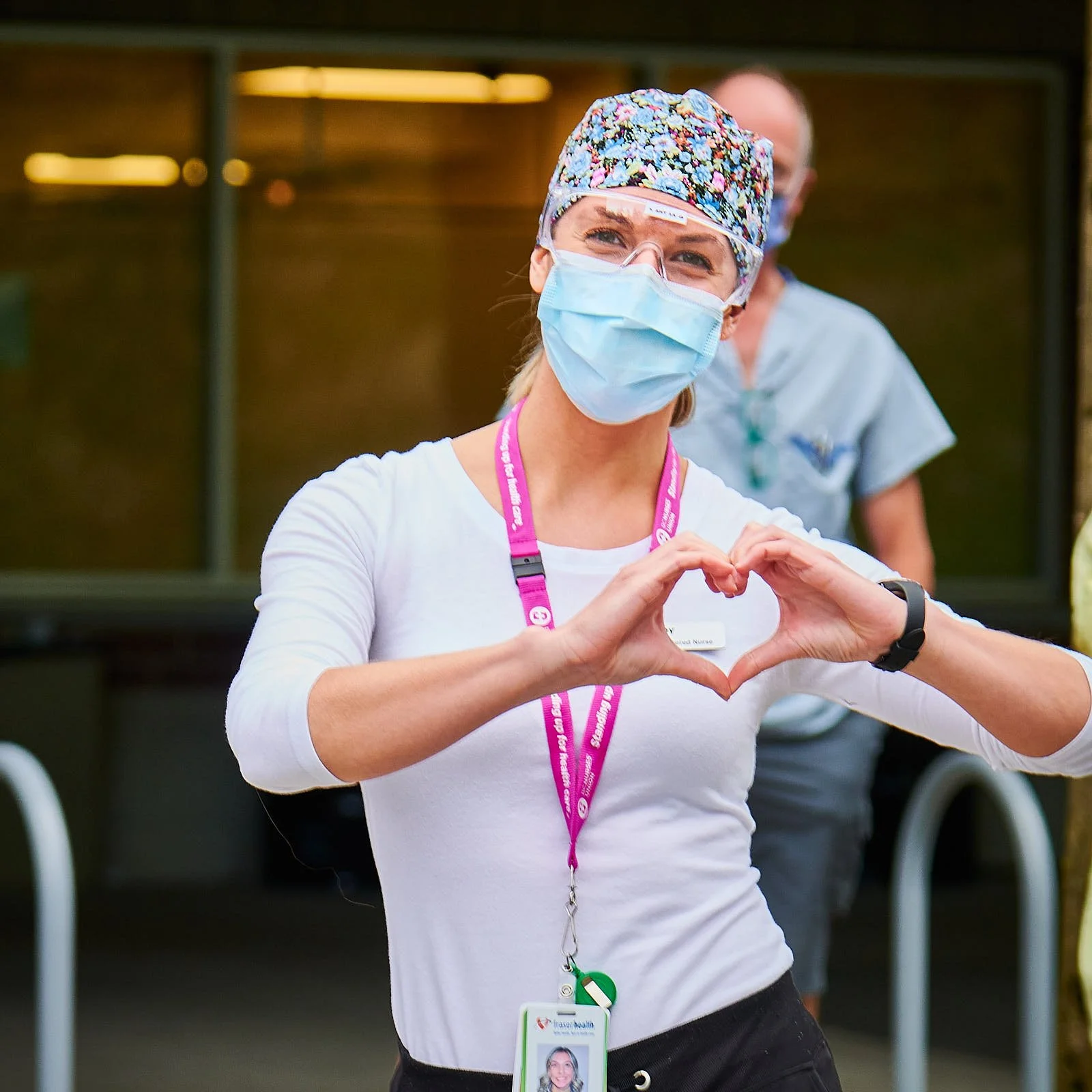 A female healthcare worker making a heart shape with her hands, wearing a face mask, colorful scrub cap, glasses, and a white shirt. Another healthcare worker is visible in the background.