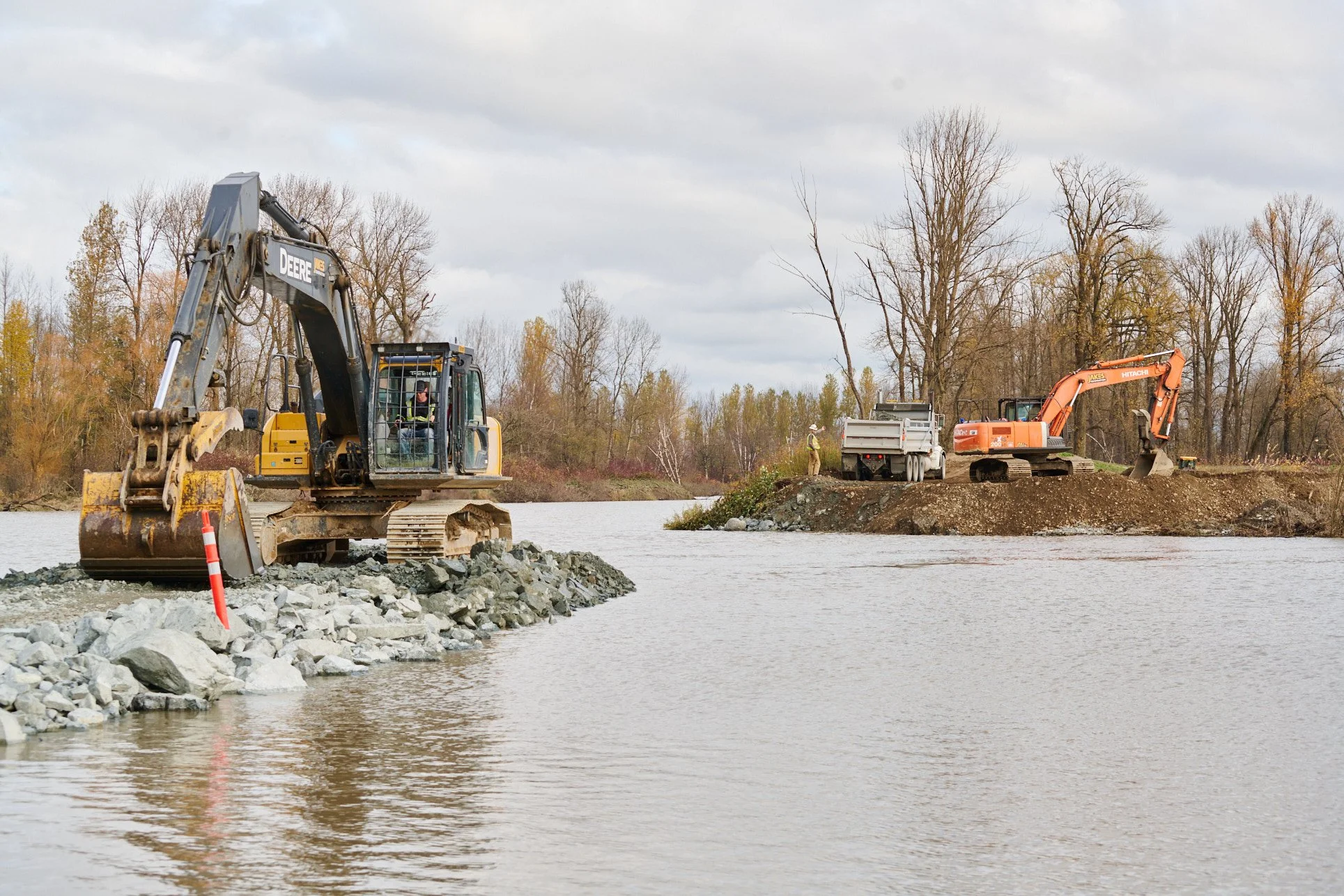 Construction site with excavators working near a river, with workers and trees in the background under cloudy sky.