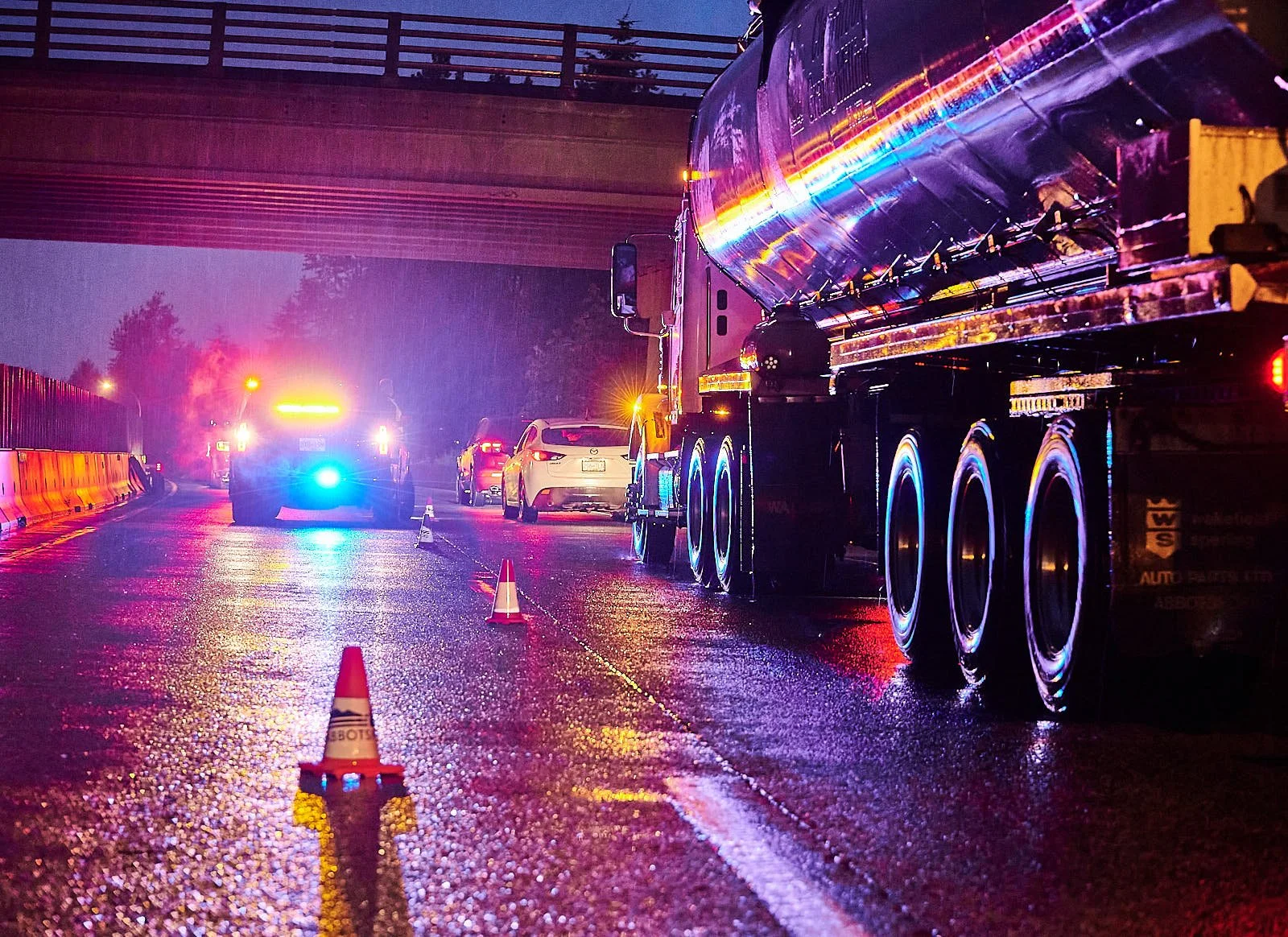 A wet road at night with emergency vehicles and cars, showing flashing lights, traffic cones, and a large truck parked on the side.