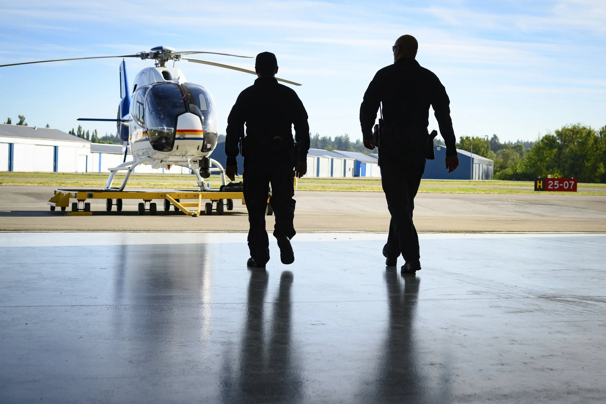 Two individuals walking towards a helicopter inside a hangar with blue skies outside.