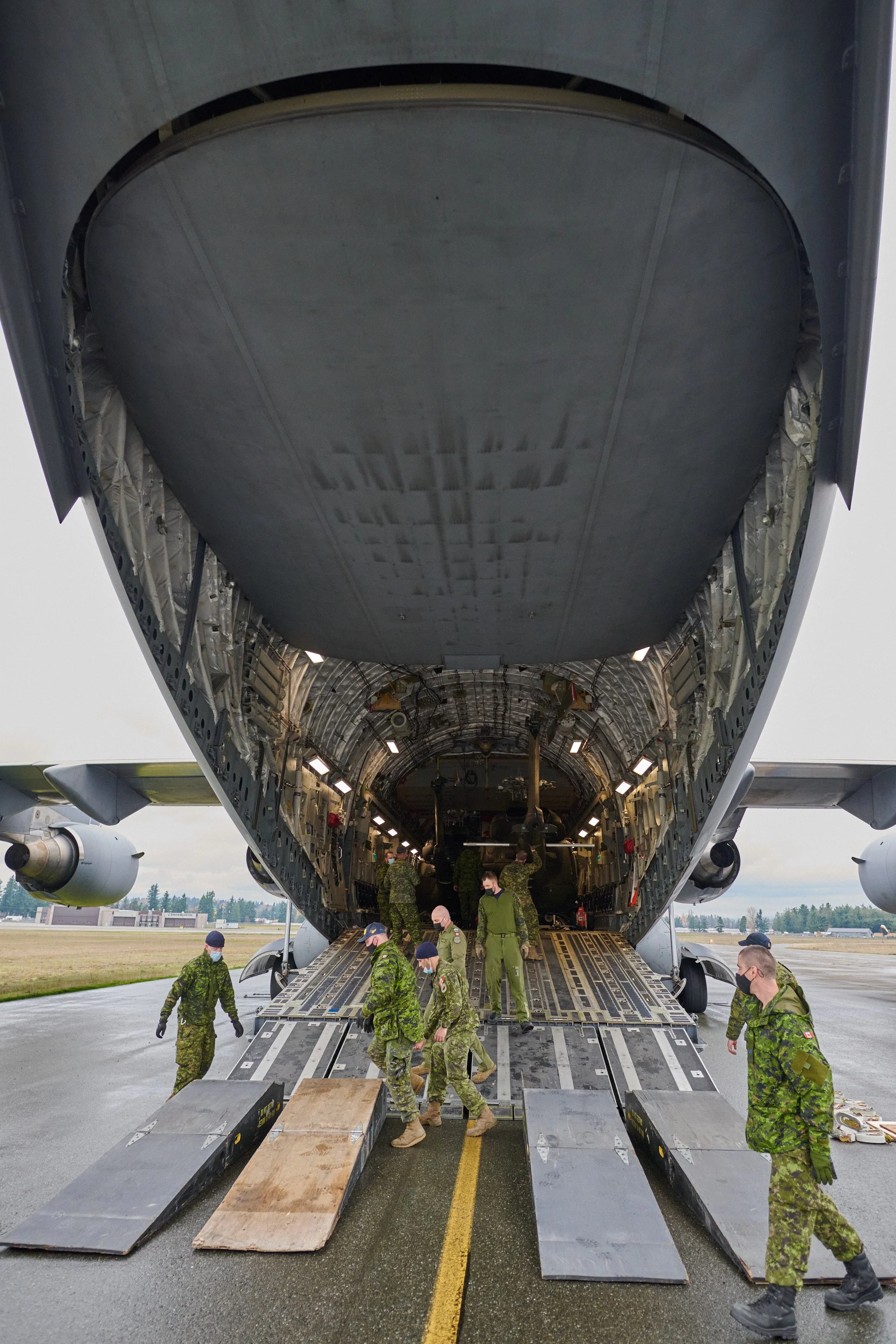 Military personnel unload supplies from the cargo hold of a large transport aircraft on an airstrip, with some soldiers standing and walking around the open rear ramp.