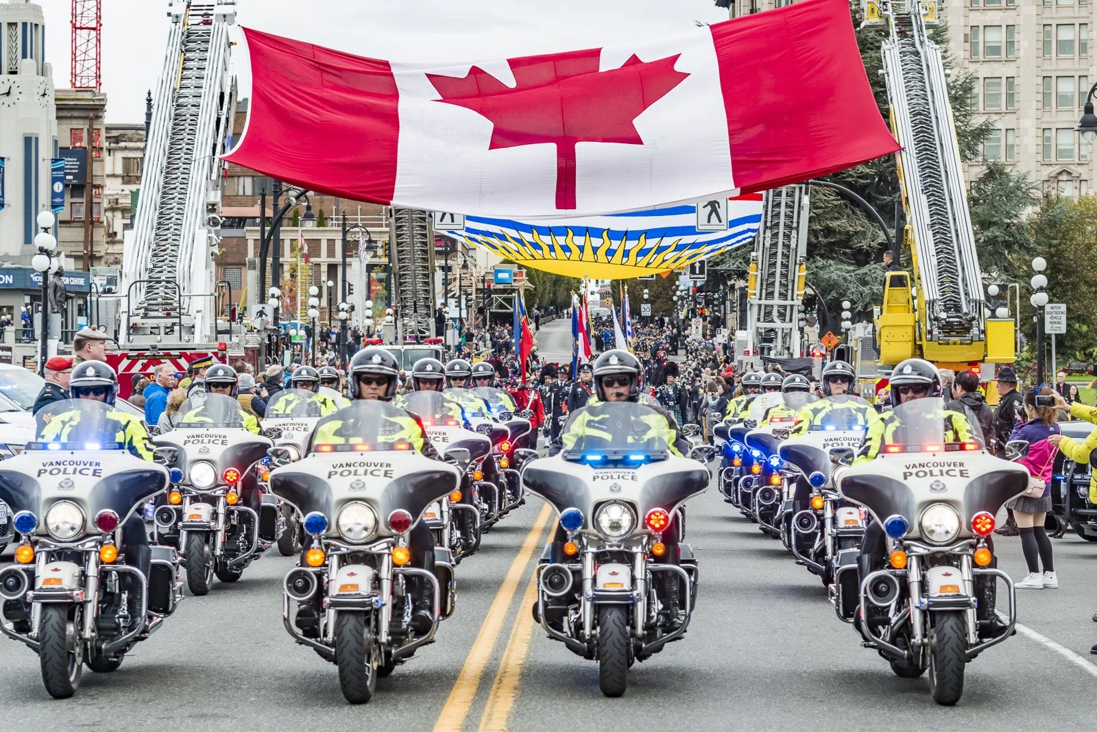 A parade of police officers on motorcycles with flashing lights is happening on a city street, with large Canadian flags hanging overhead and a crowd of spectators lining the sides of the street.