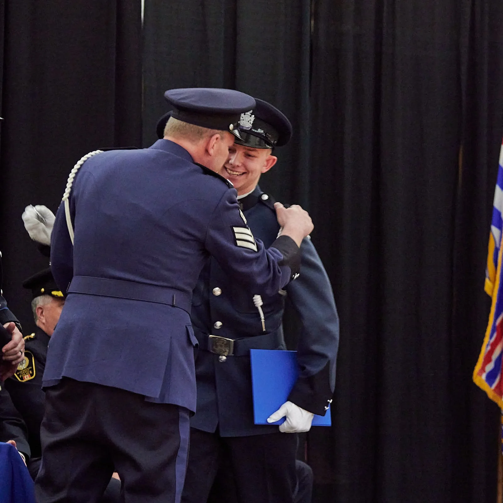 A uniformed police officer receiving an award or recognition from another officer in a blue uniform, with other officers and a black curtain in the background.