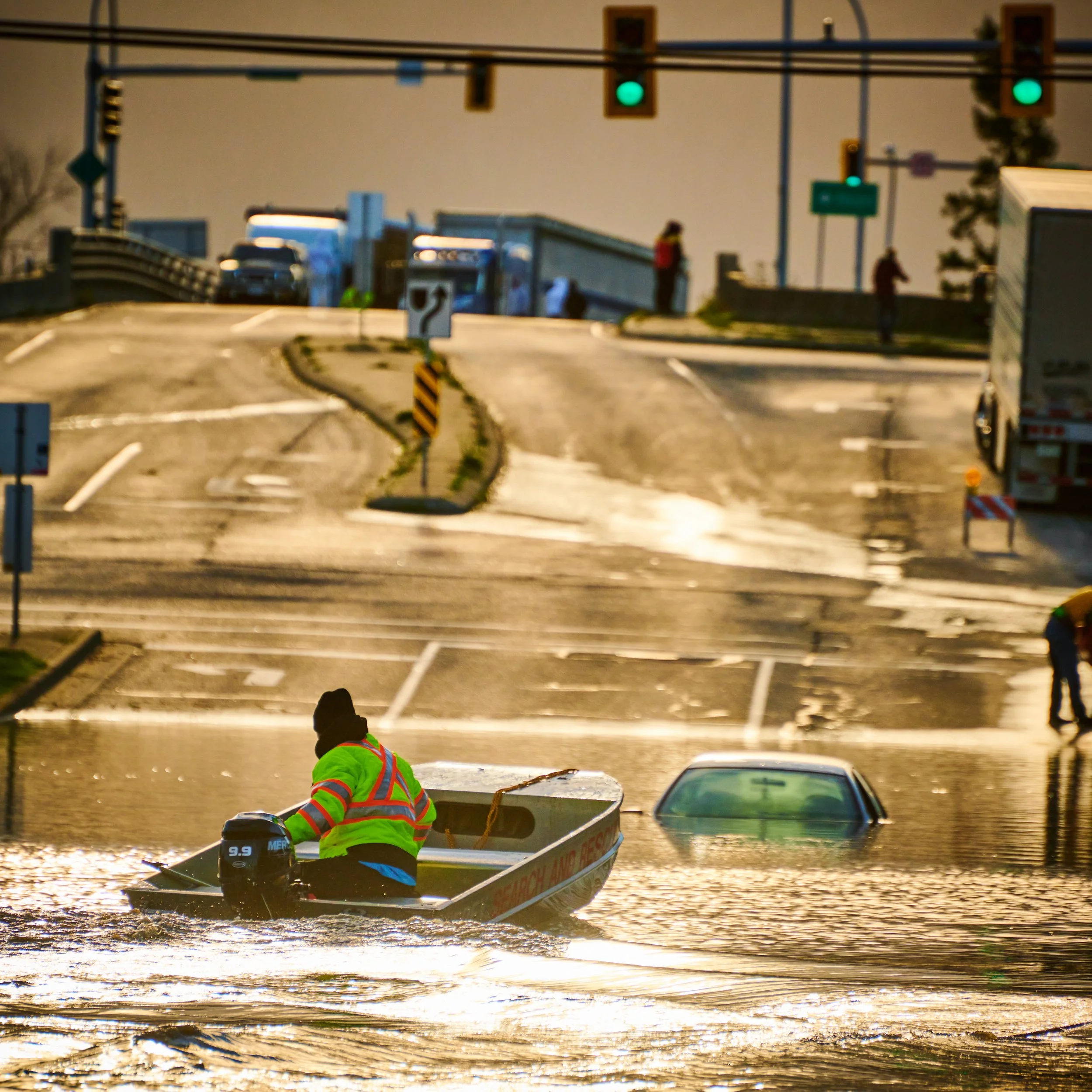 A person in a bright green safety vest and helmet paddles a small motorized boat through floodwaters with submerged road markings and traffic signals in the background.