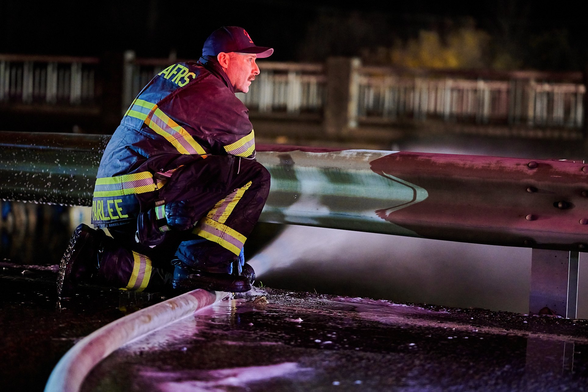 Firefighter kneeling on wet ground near a fire truck at night, holding a hose, with a guardrail and blurred trees in the background.