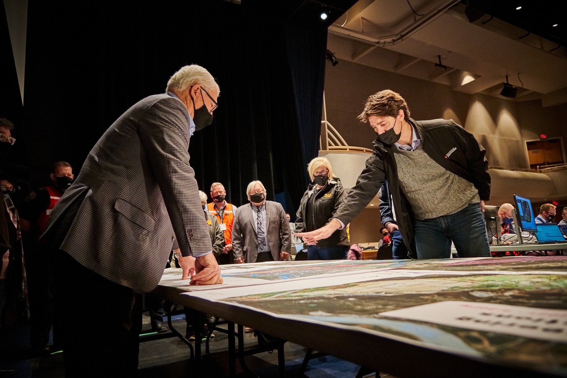People gathered around a table examining maps or plans at an indoor event. Most individuals are wearing masks, and some are looking at the table while others observe.
