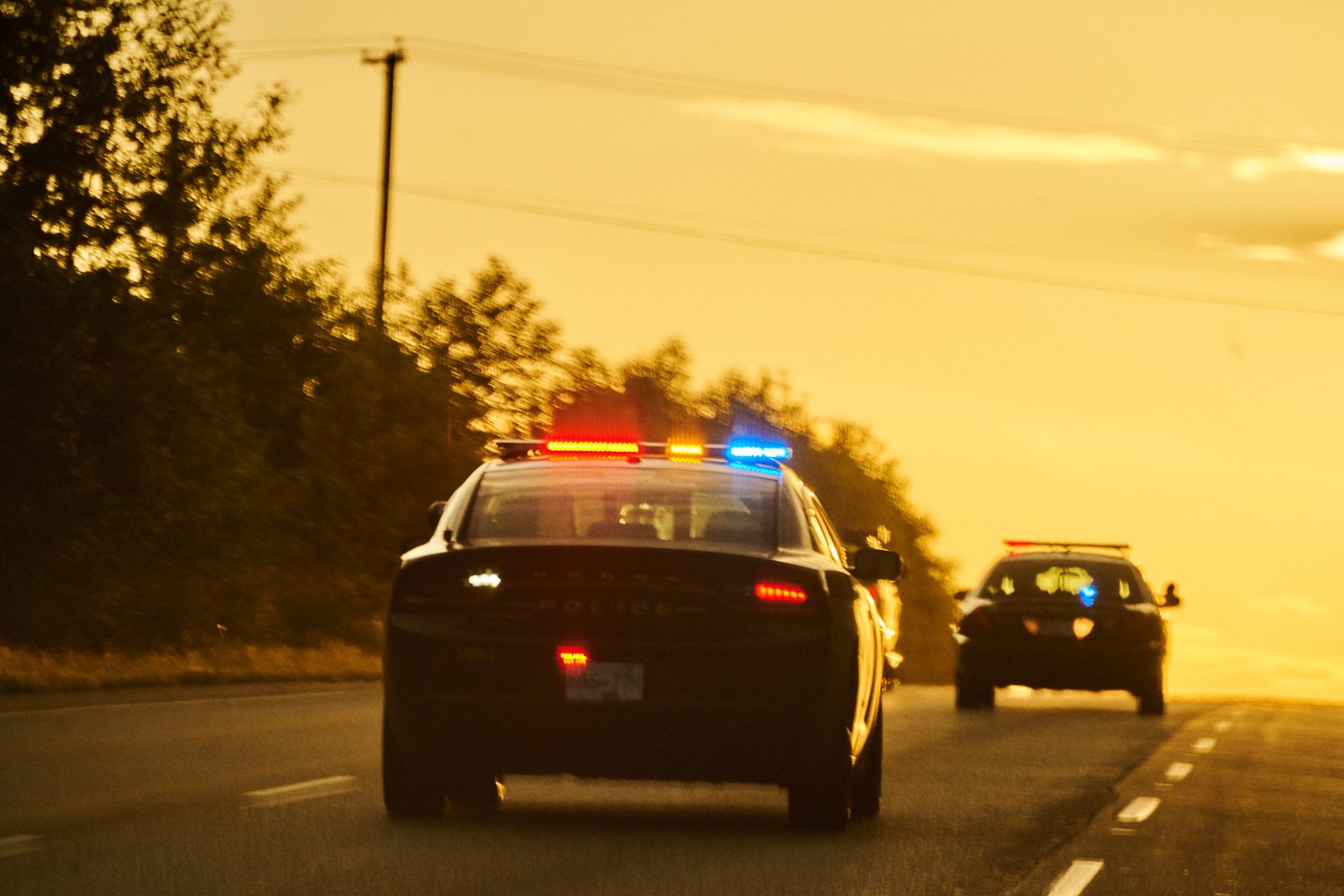 Two police cars with flashing red, blue, and yellow lights on a highway during sunset.