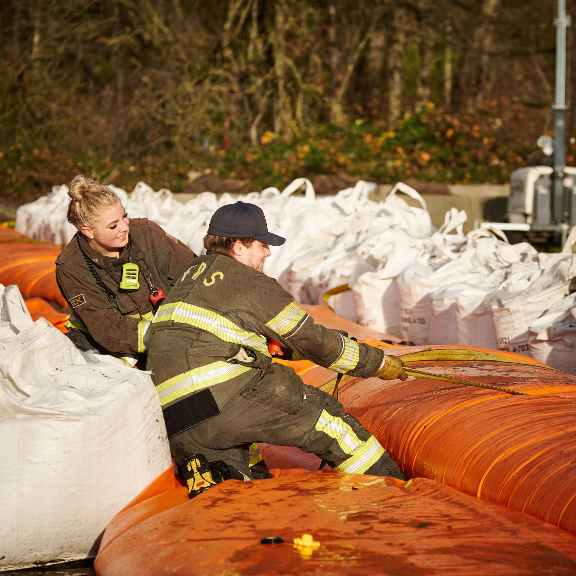 Firefighters working together at an outdoor scene during daytime, with large white bags and orange equipment visible.
