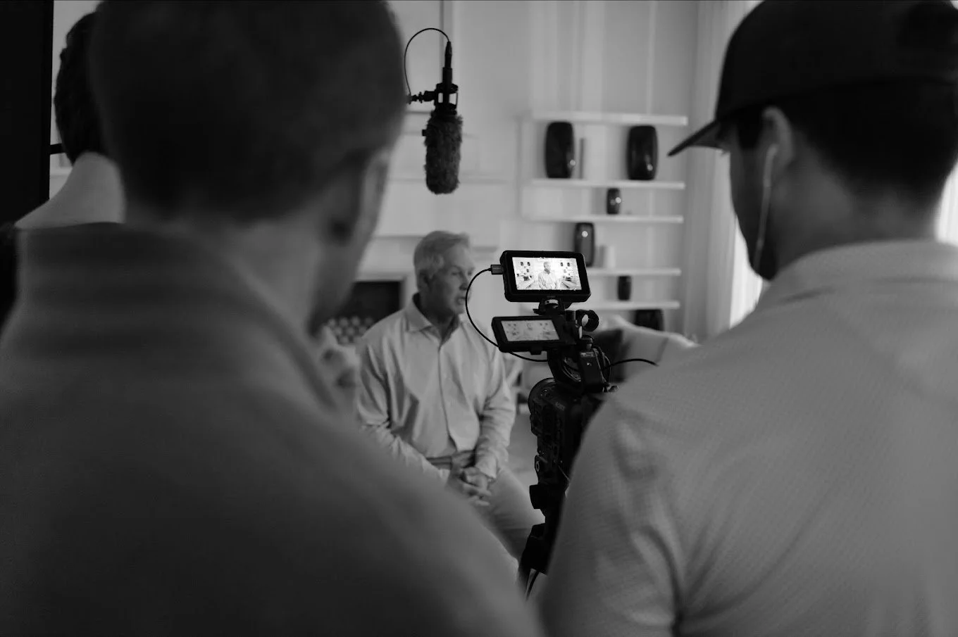 People filming a man sitting in a living room for a video or interview, with professional filming equipment and a camera.
