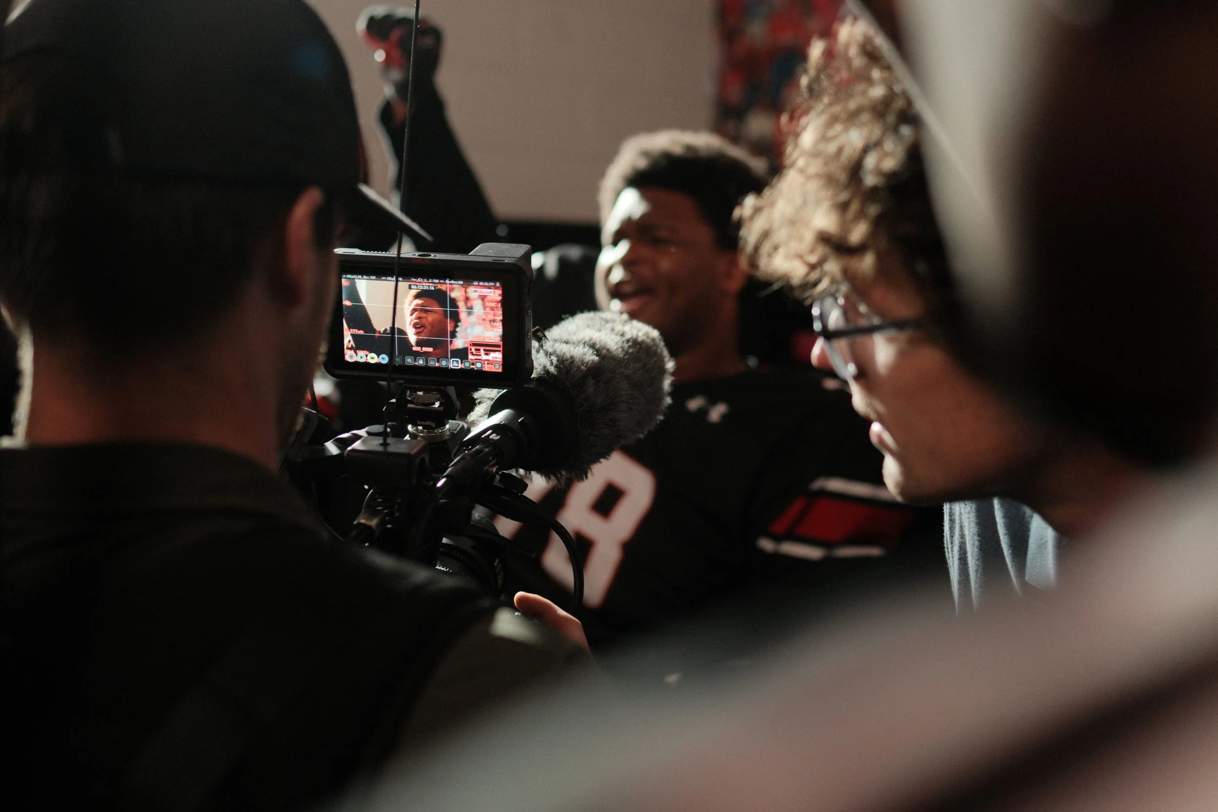 A person is filming or photographing a young man wearing a football jersey, who is smiling and engaged in conversation or an interview. The scene appears to be indoors with a dark background.