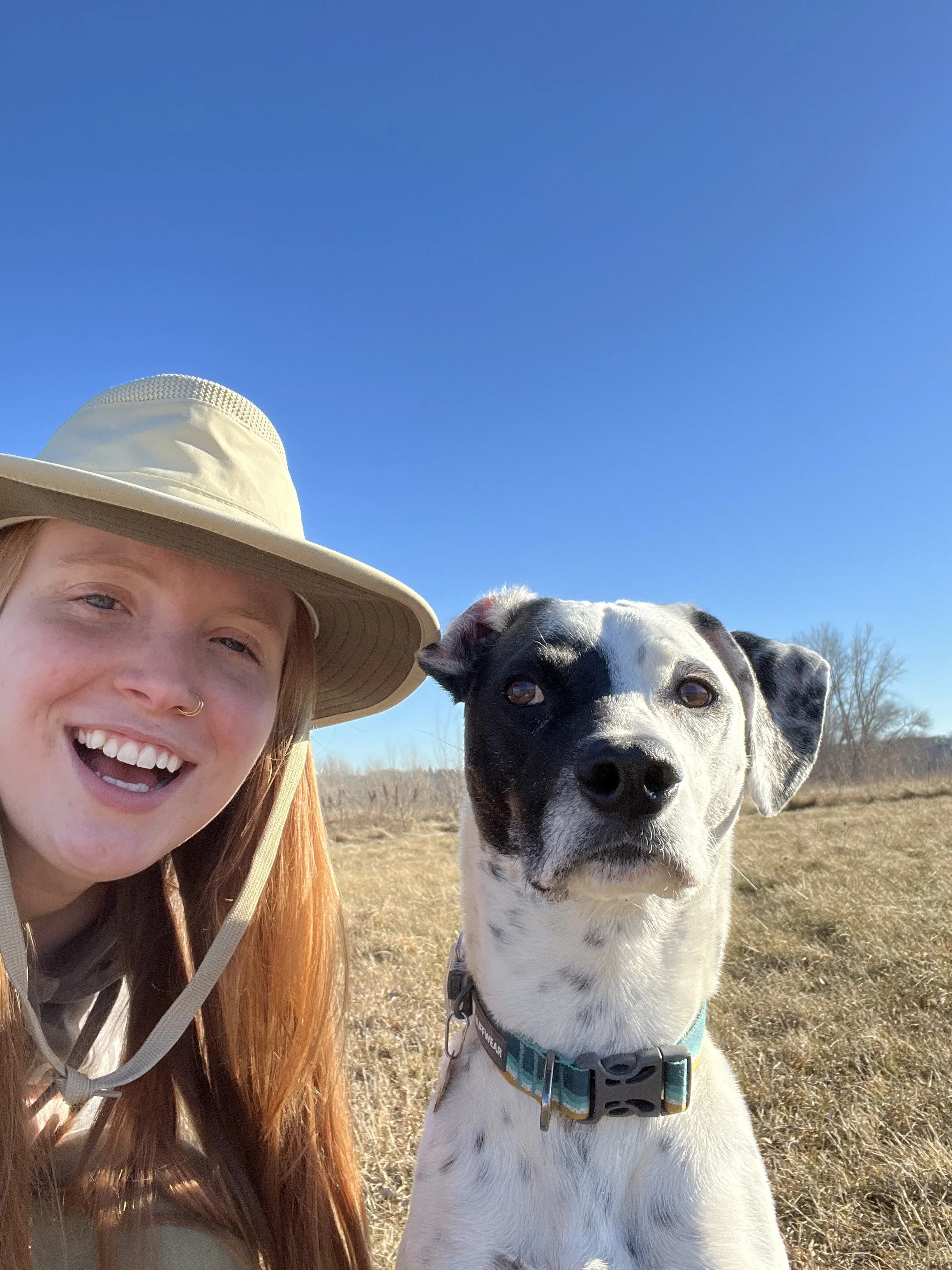 Lily, a white femme person with long red hair, smiles beside their medium-sized, white with black spotted dog.