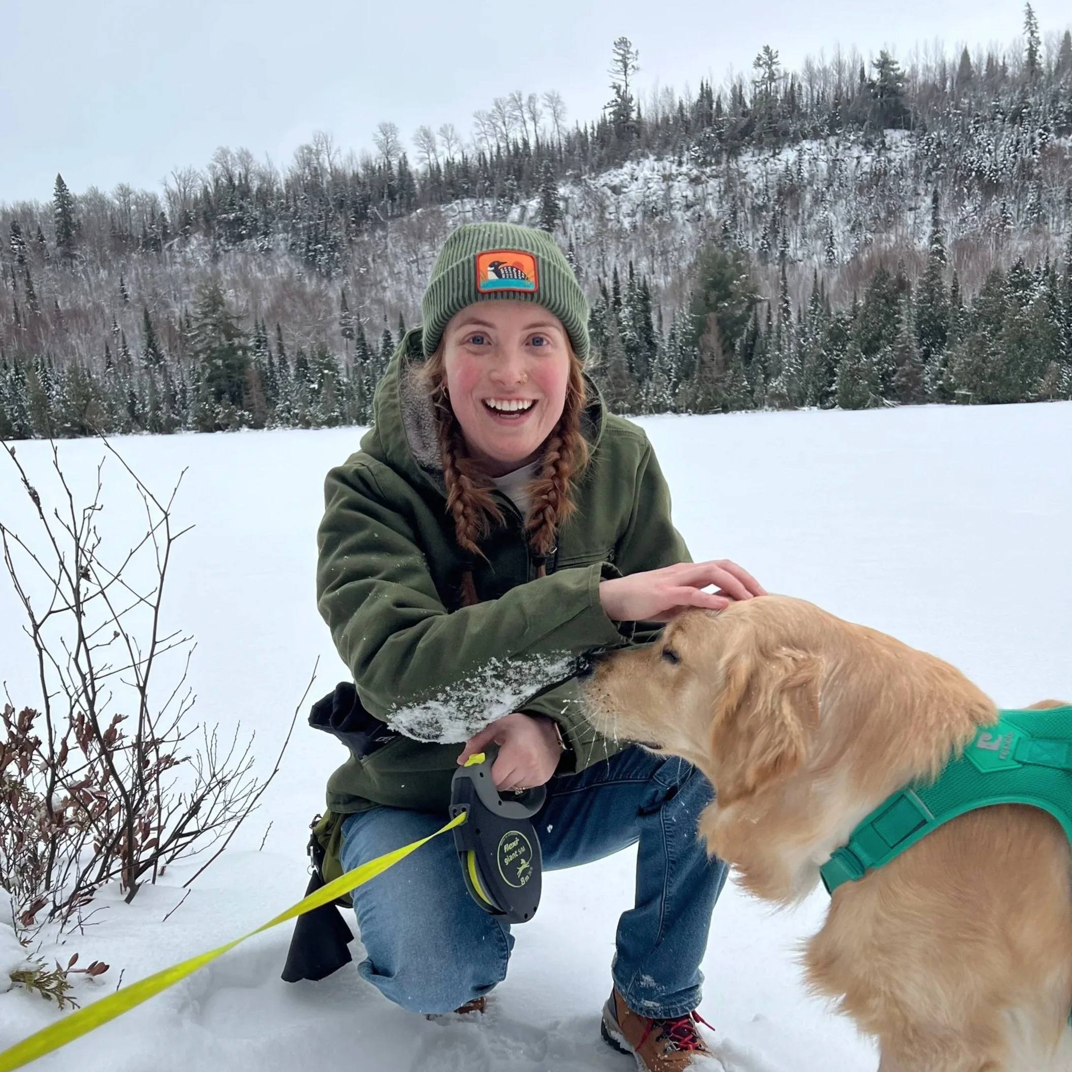 Lily smiles and kneels in the snow next to a golden, scratching his head