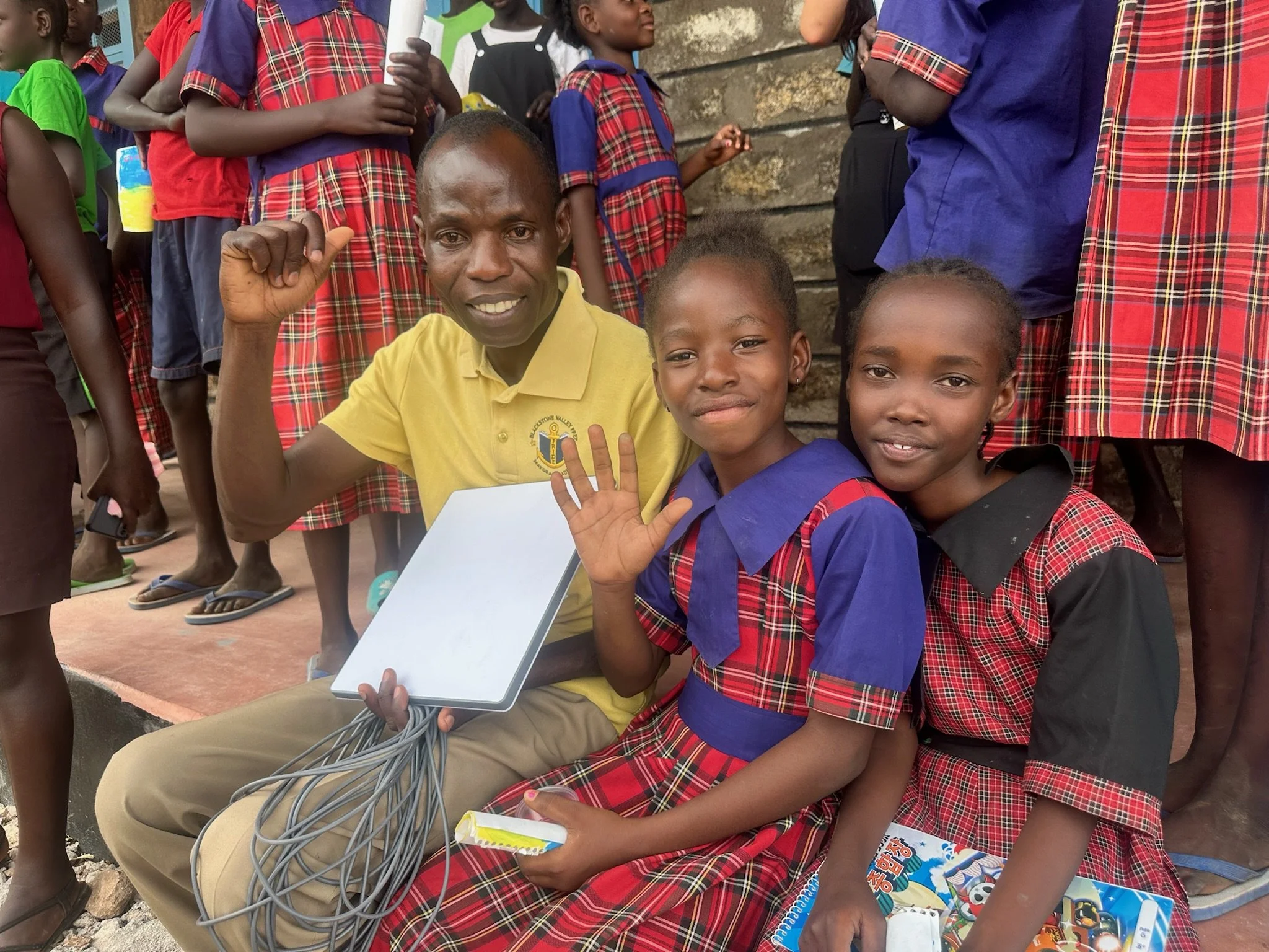two schools children and a teacher sitting and holding a starlink device and smiling while waving