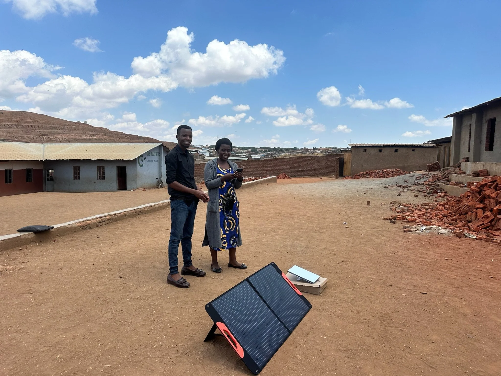 two school teachers in malawi smiling as they stand in front of a solar panel and a starlink device