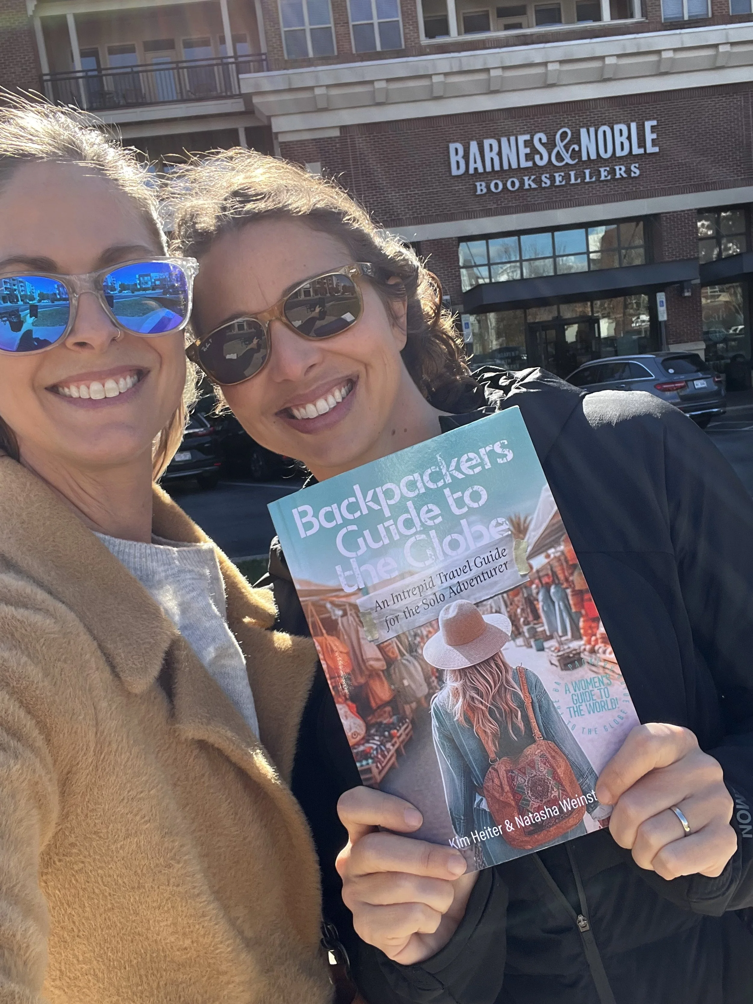 Two women holding their book wearing sunglasses. The woman on the left is blonde wearing a brown teddy bear jacket, the woman on right is brunette wearing black jacket. Both standing in front of a Barnes & Noble bookstore.