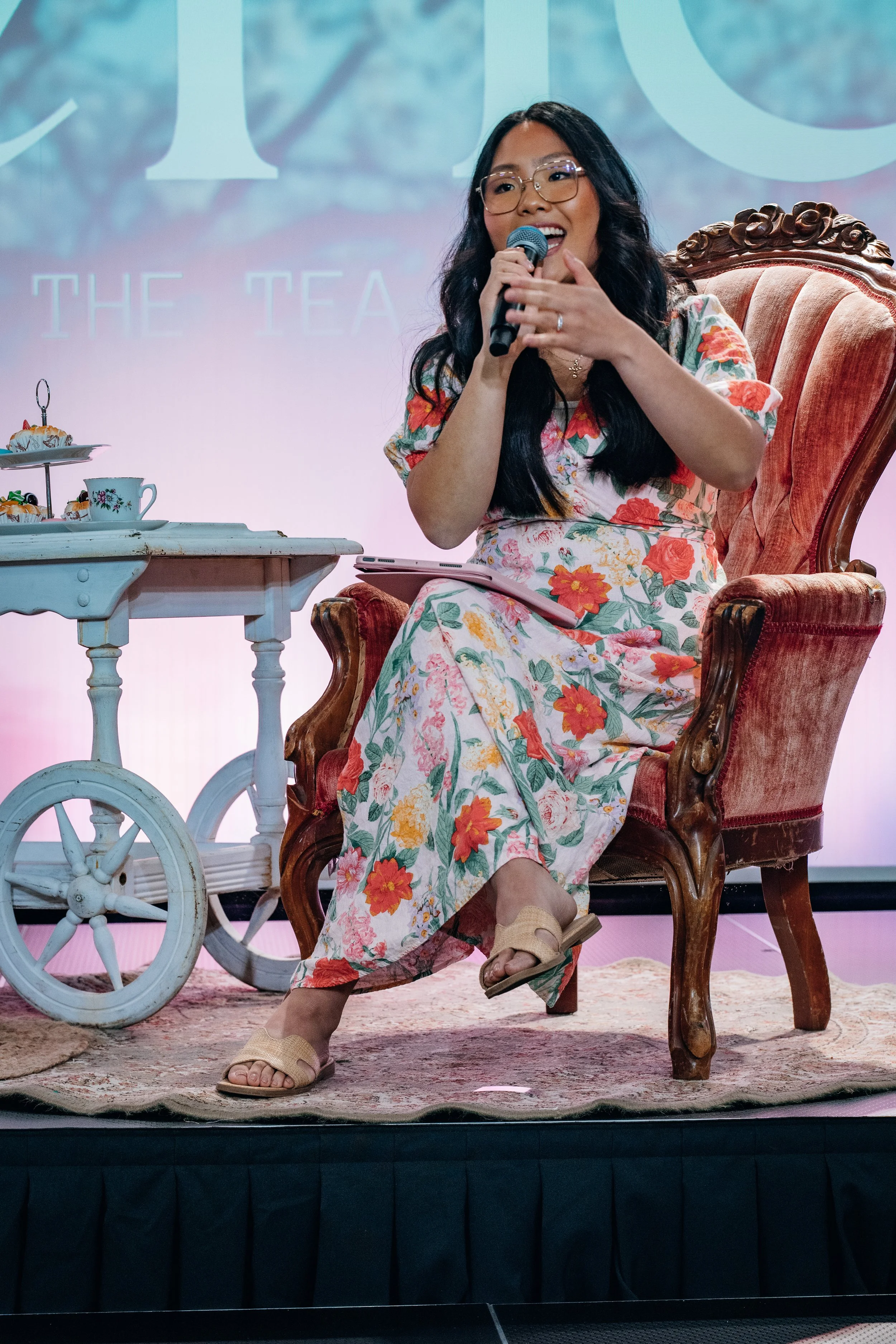 Tara in a floral dress speaking into a microphone while seated on a vintage chair at a women’s event, with a tea cart beside her on stage.