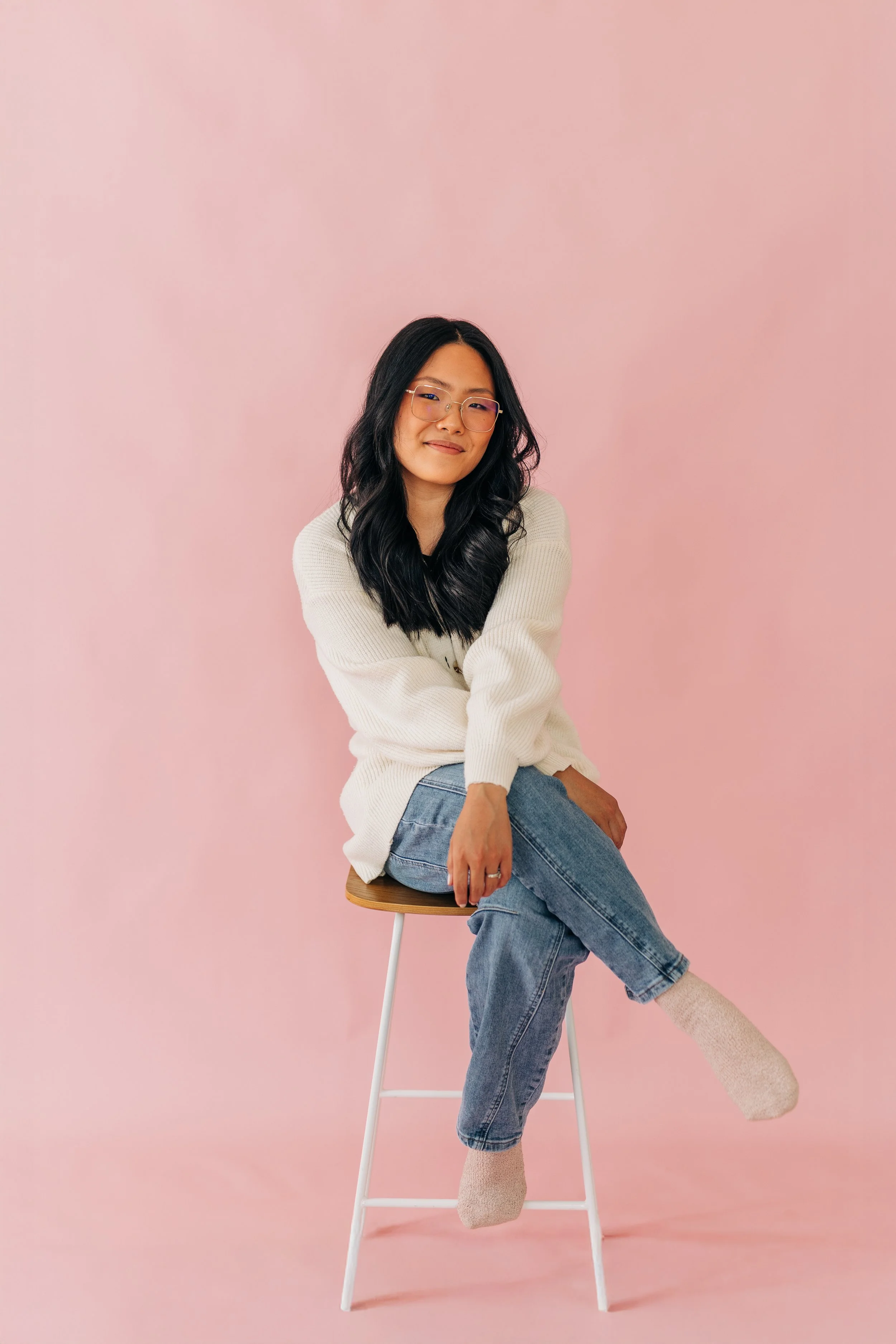 Tara wearing a cream sweater and jeans sitting on a stool against a soft pink background, smiling gently at the camera.