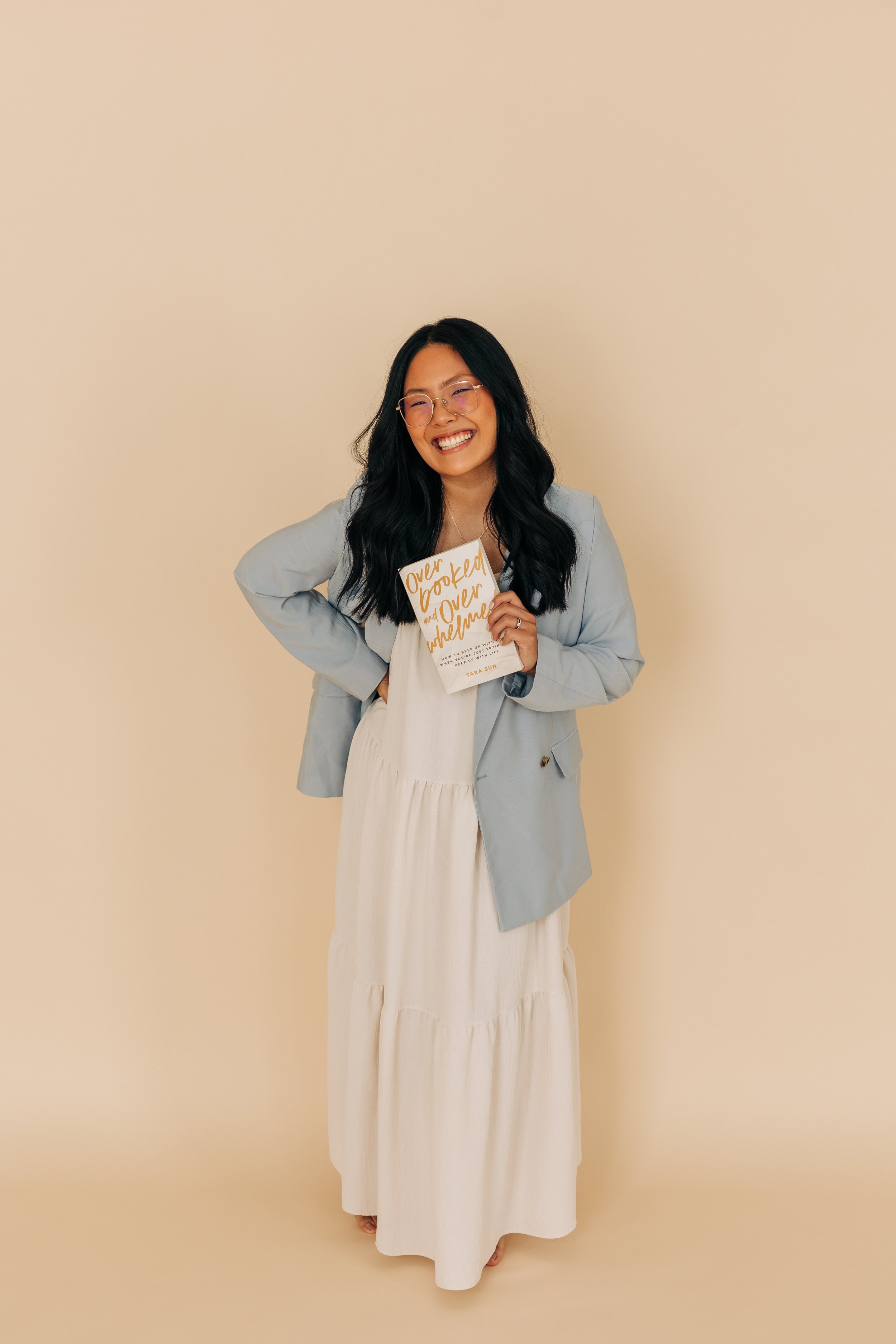 Tara smiling in a light blue blazer and white dress holding her book Overlooked and Overwhelmed against a neutral beige background.