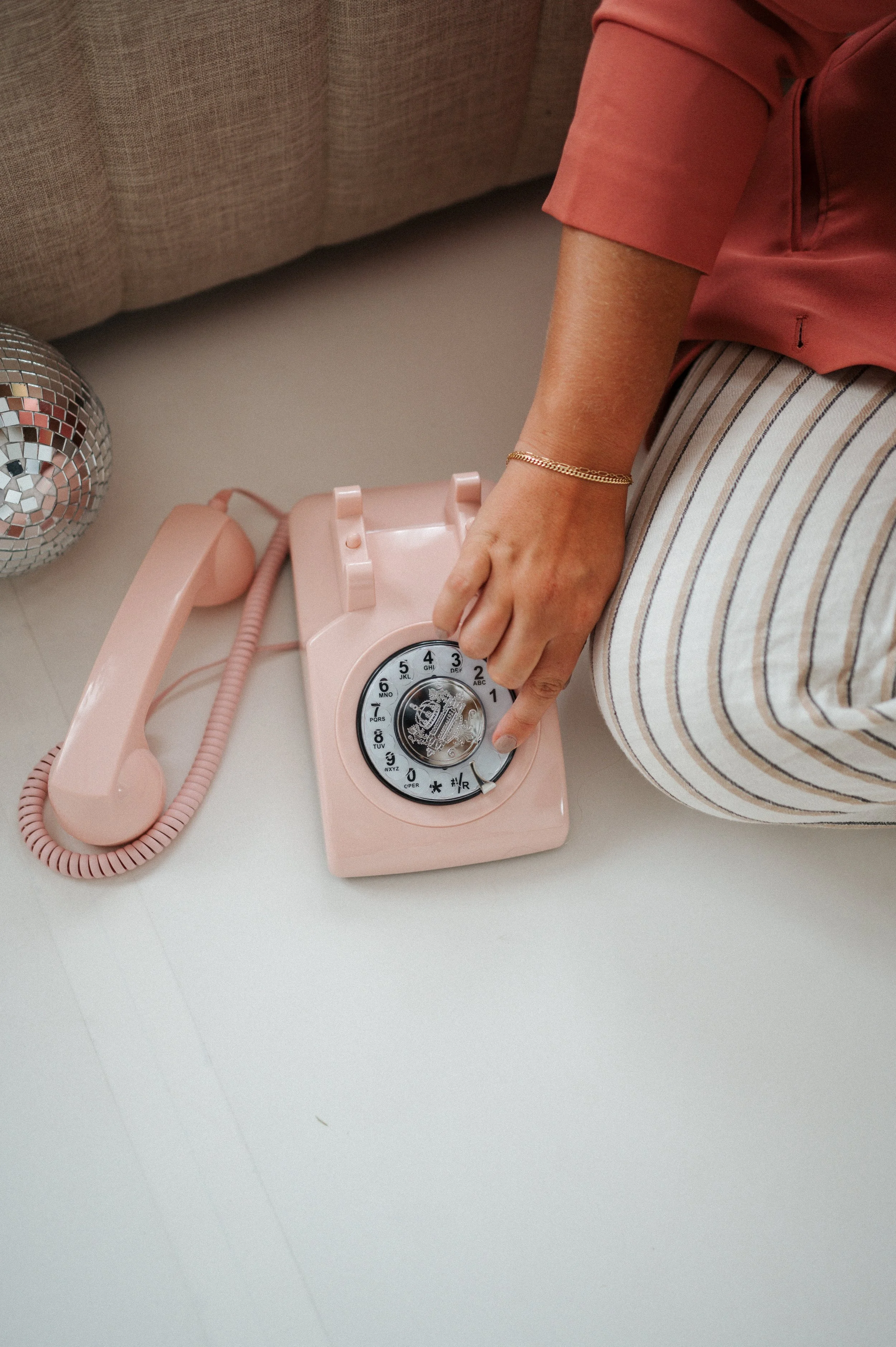 Hand dialing a pastel pink rotary phone in a feminine, retro-inspired flat lay with a disco ball and soft striped fabric.
