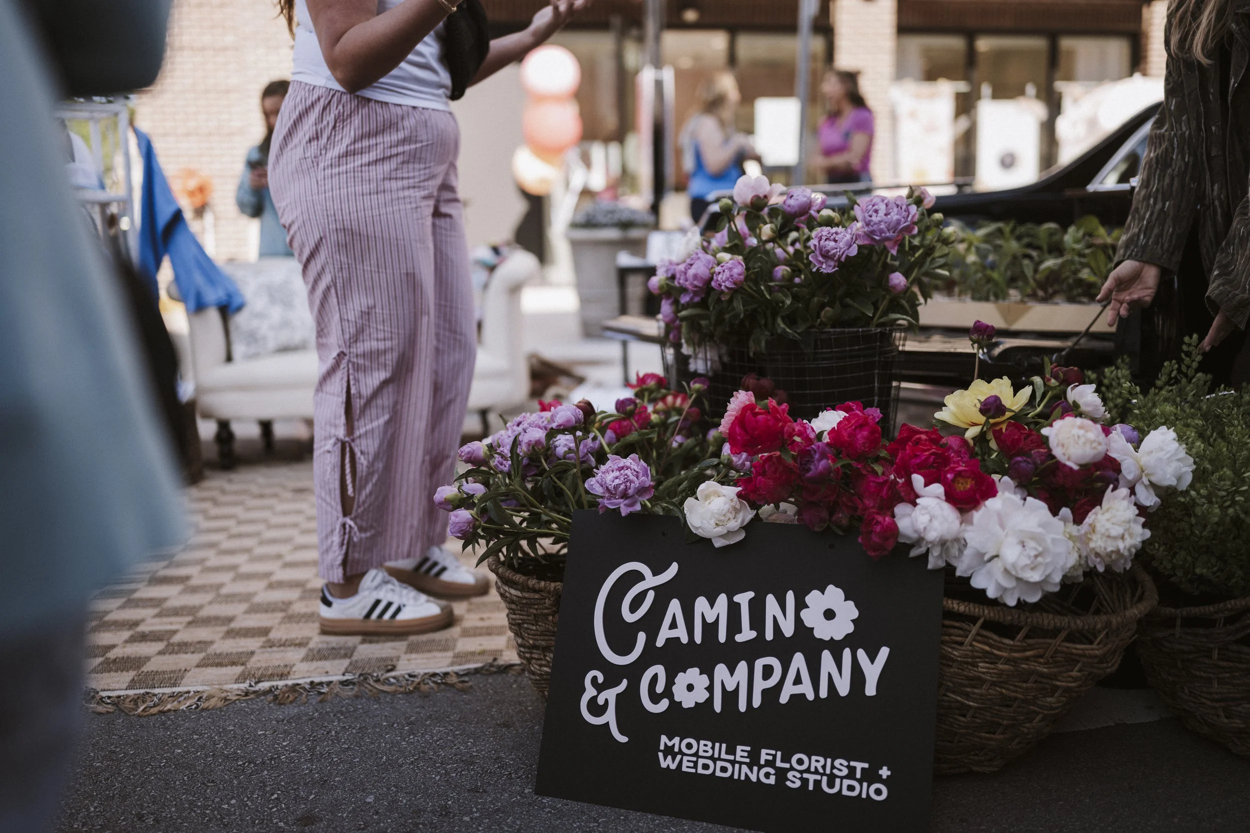Camino & Company branded market display with peony-filled flower baskets and mobile florist signage at an outdoor event.