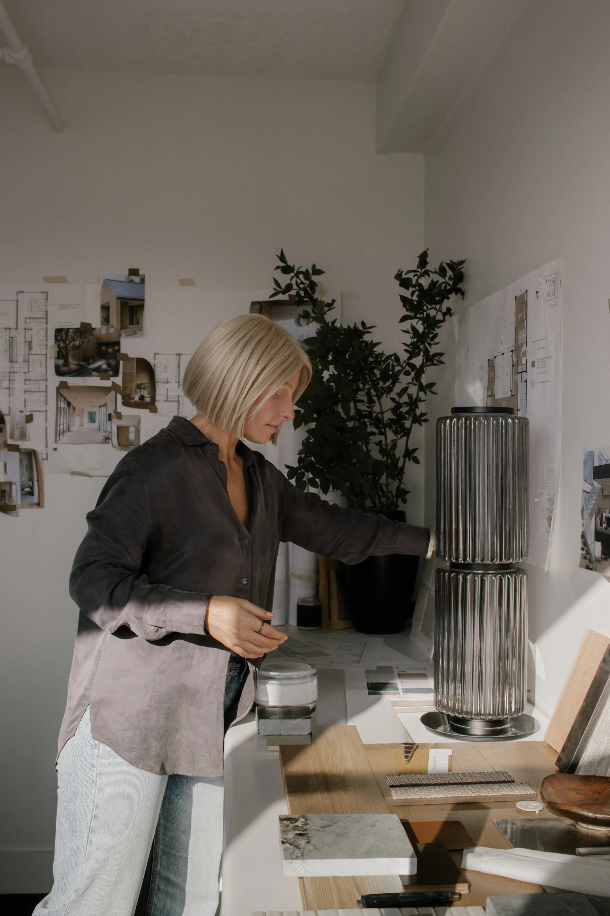 Woman with blonde hair in a business casual outfit viewing design material in a modern workspace with interior design plans on the wall and decorative items on the table.