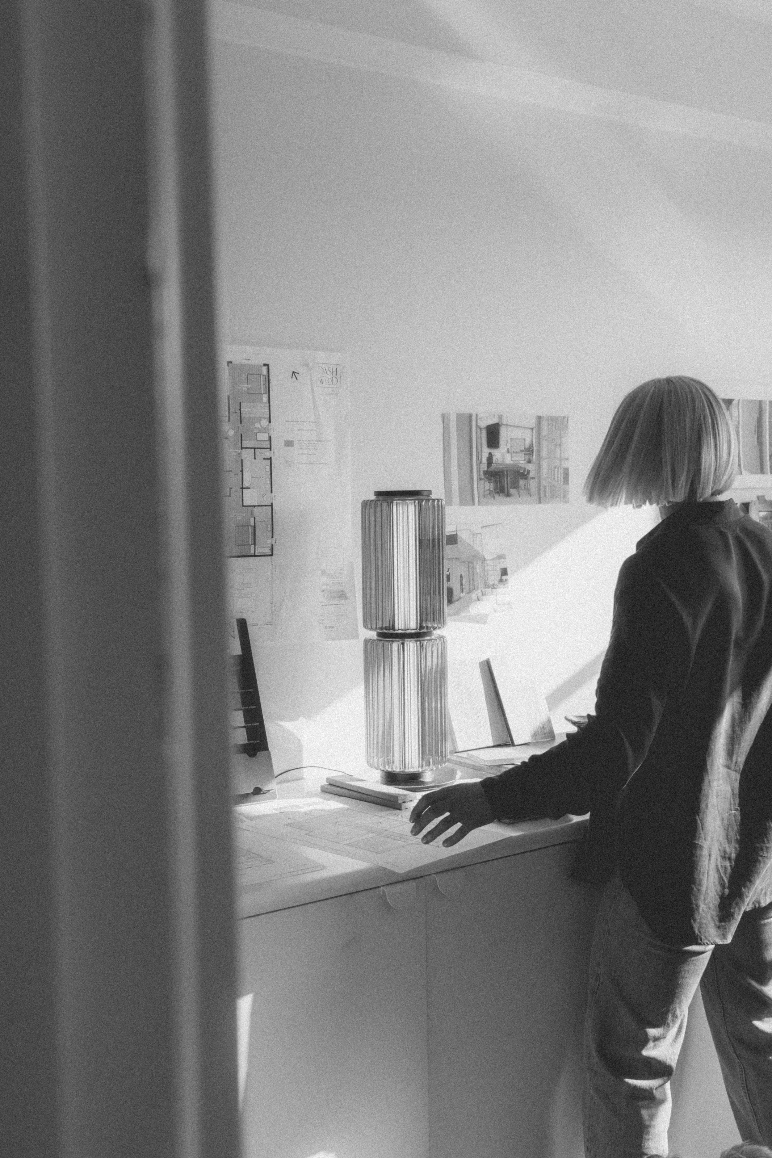 A woman with shoulder-length hair stands in front of a desk with architectural drawings and a lamp, viewed from behind. The scene appears to be in an office or studio with various sketches and plans posted on the wall.