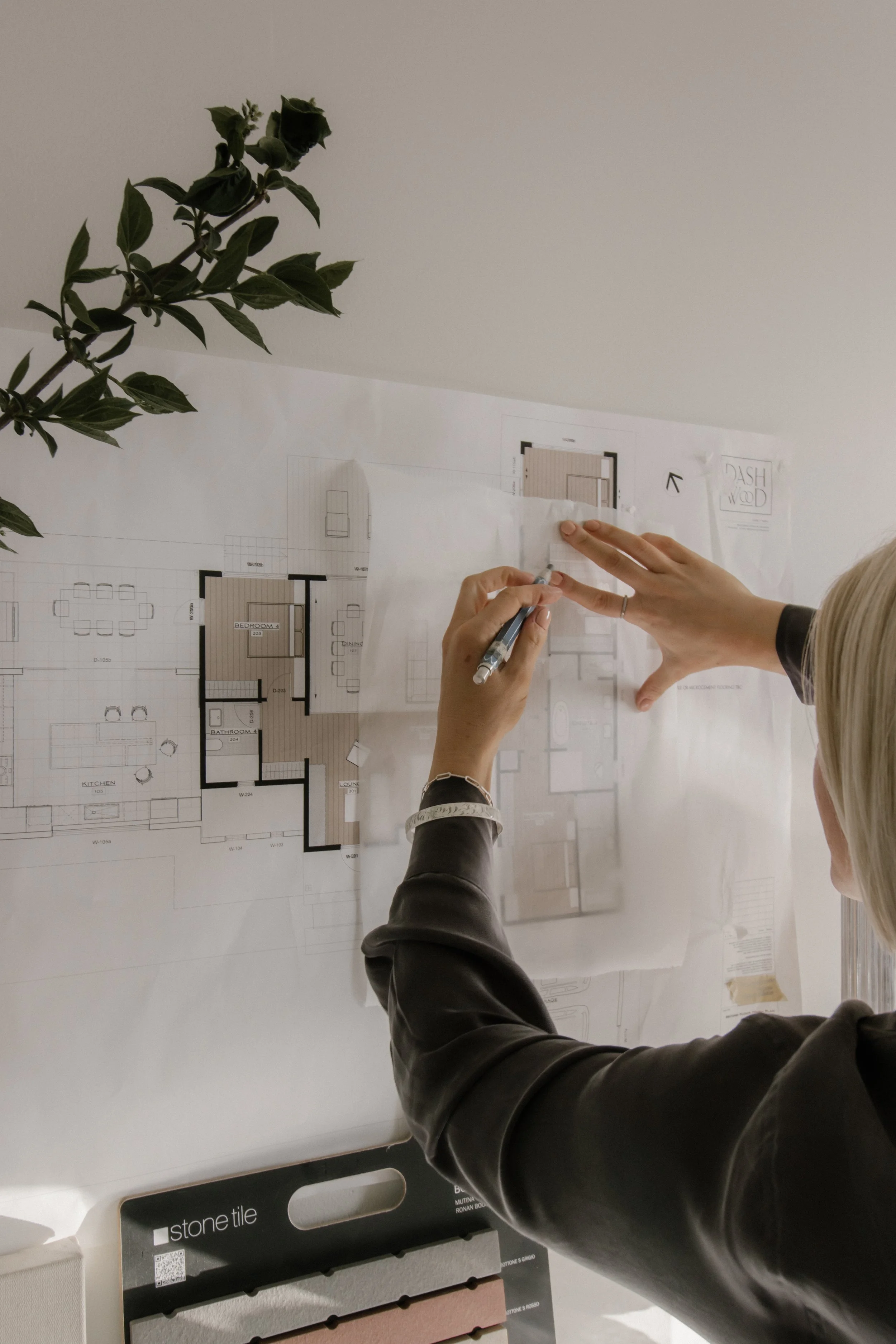 Person in a black leather jacket using a pen to point at a blueprint or floor plan on a table. A potted plant and samples of stone tile materials are visible on the table.