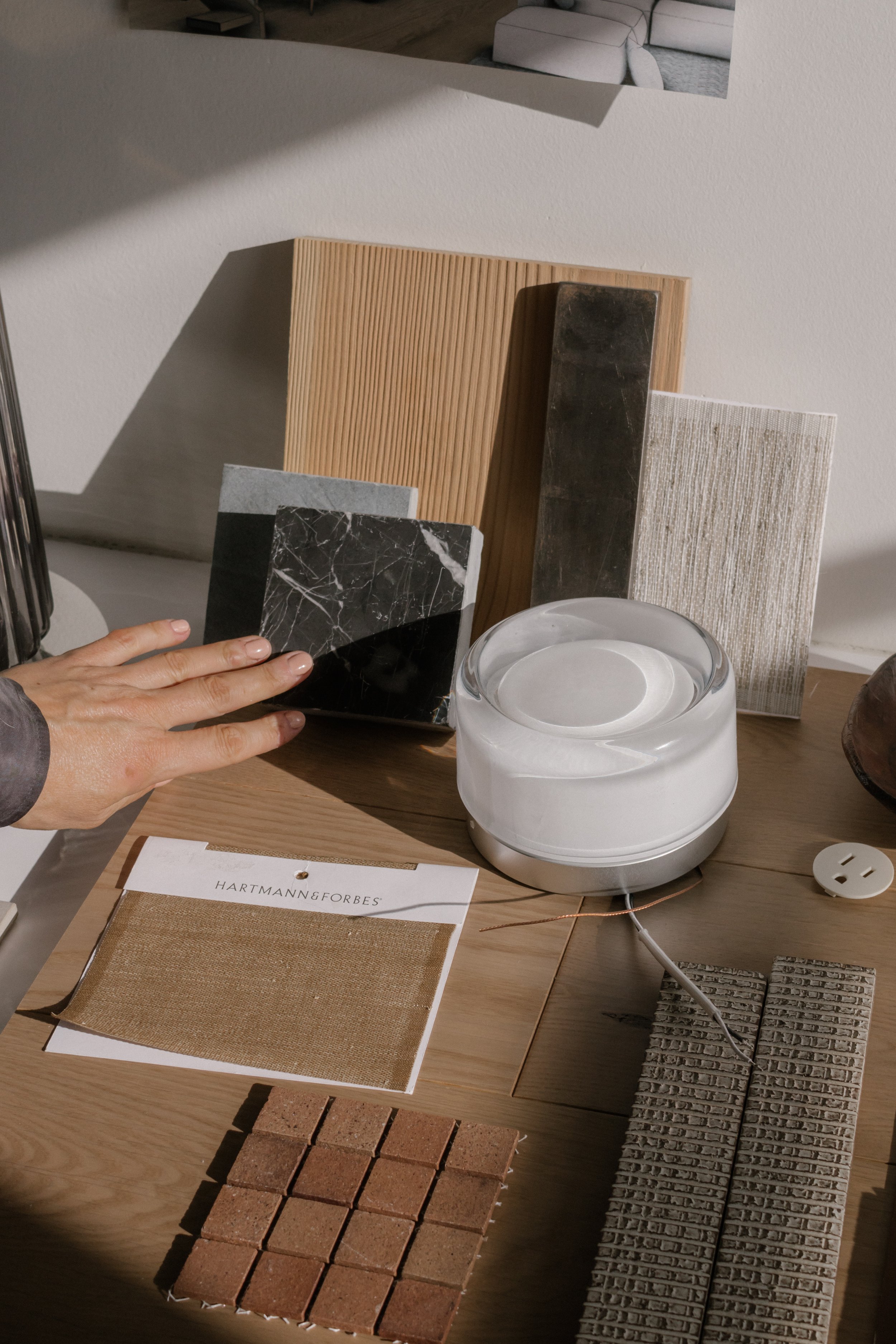 A variety of tile samples, fabric swatch, color palette, and a sample of marble are arranged on a wooden surface, with a scale and a circular glass container nearby.