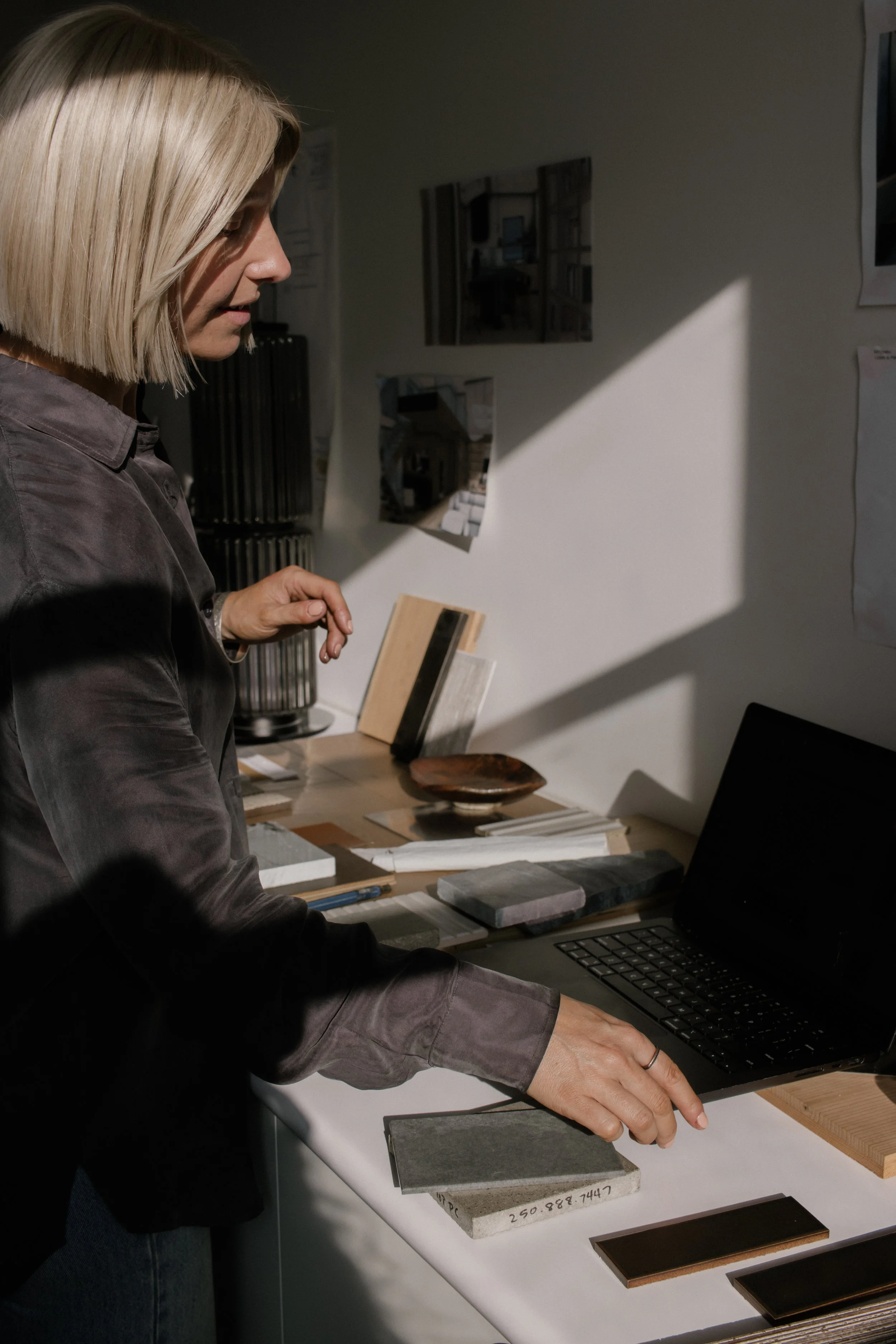Woman with blonde hair in a bob cut looking at design samples on a table, with a laptop in front of her, in an art studio or design workspace.