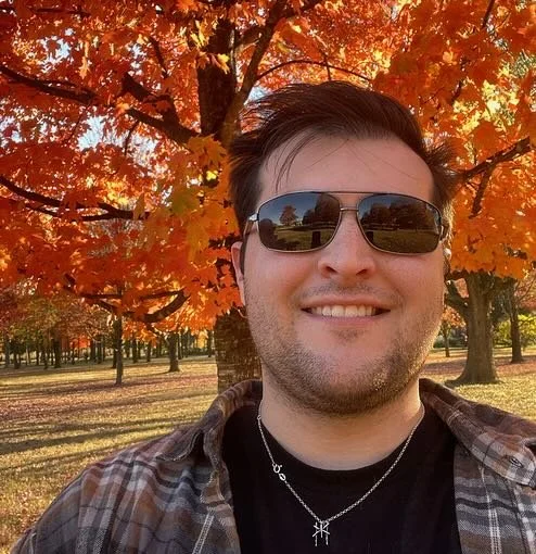 A man smiling outdoors in front of a tree with orange fall foliage, wearing sunglasses and a plaid shirt.