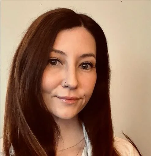 A woman with long brown hair, wearing a nose ring, smiling slightly, against a plain background.
