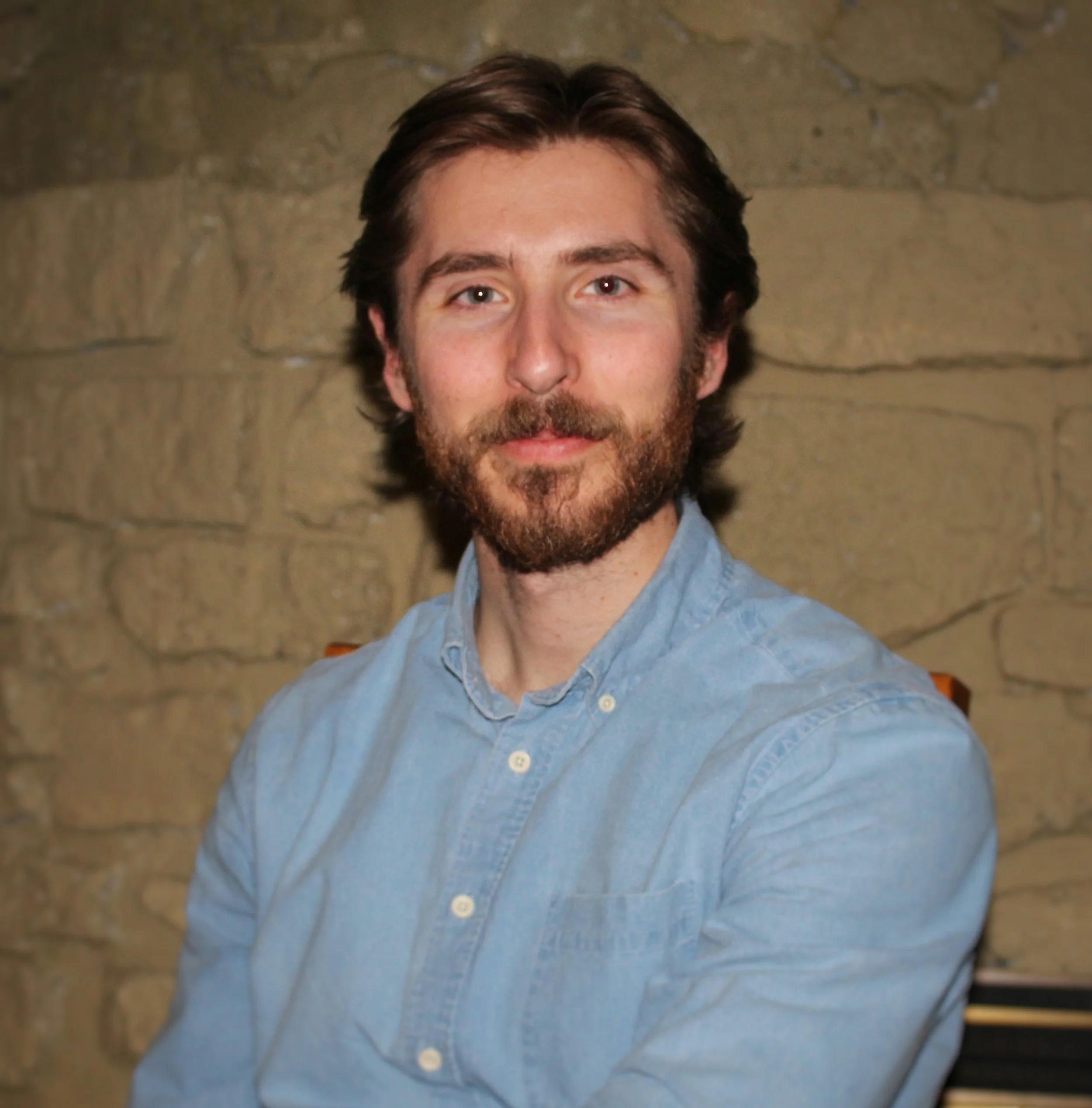 A man with dark brown hair, a beard, and blue eyes, wearing a light blue button-up shirt, is sitting in front of a stone wall.
