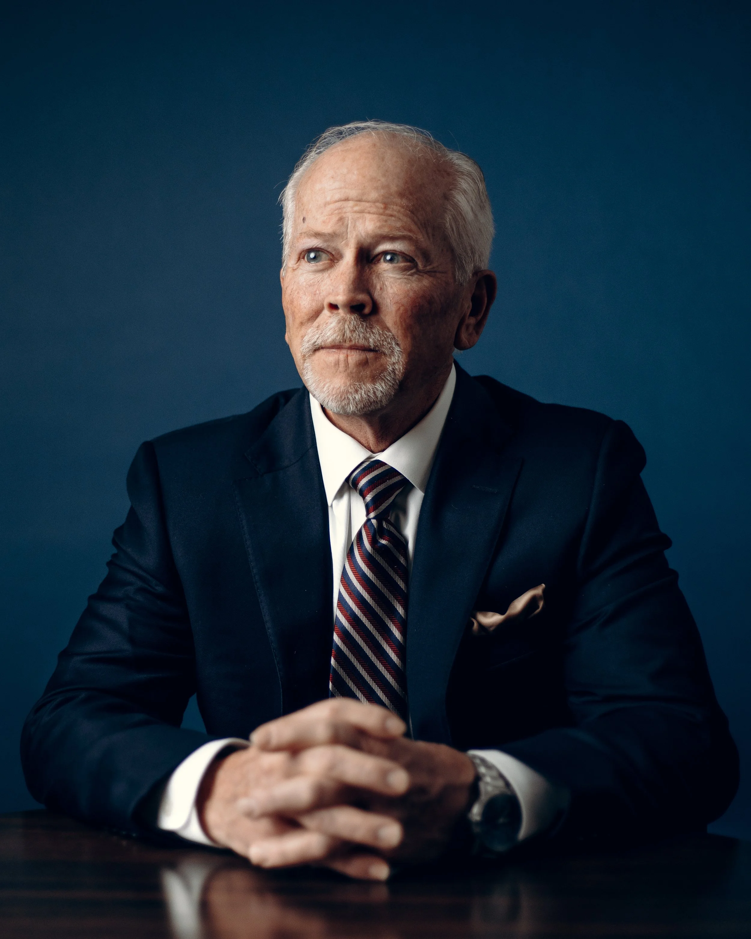 Elderly man in suit with striped tie sitting at a table with hands clasped.