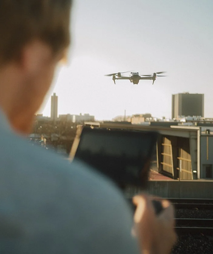 A person is watching a drone fly over a cityscape from a rooftop, holding a remote control or device.