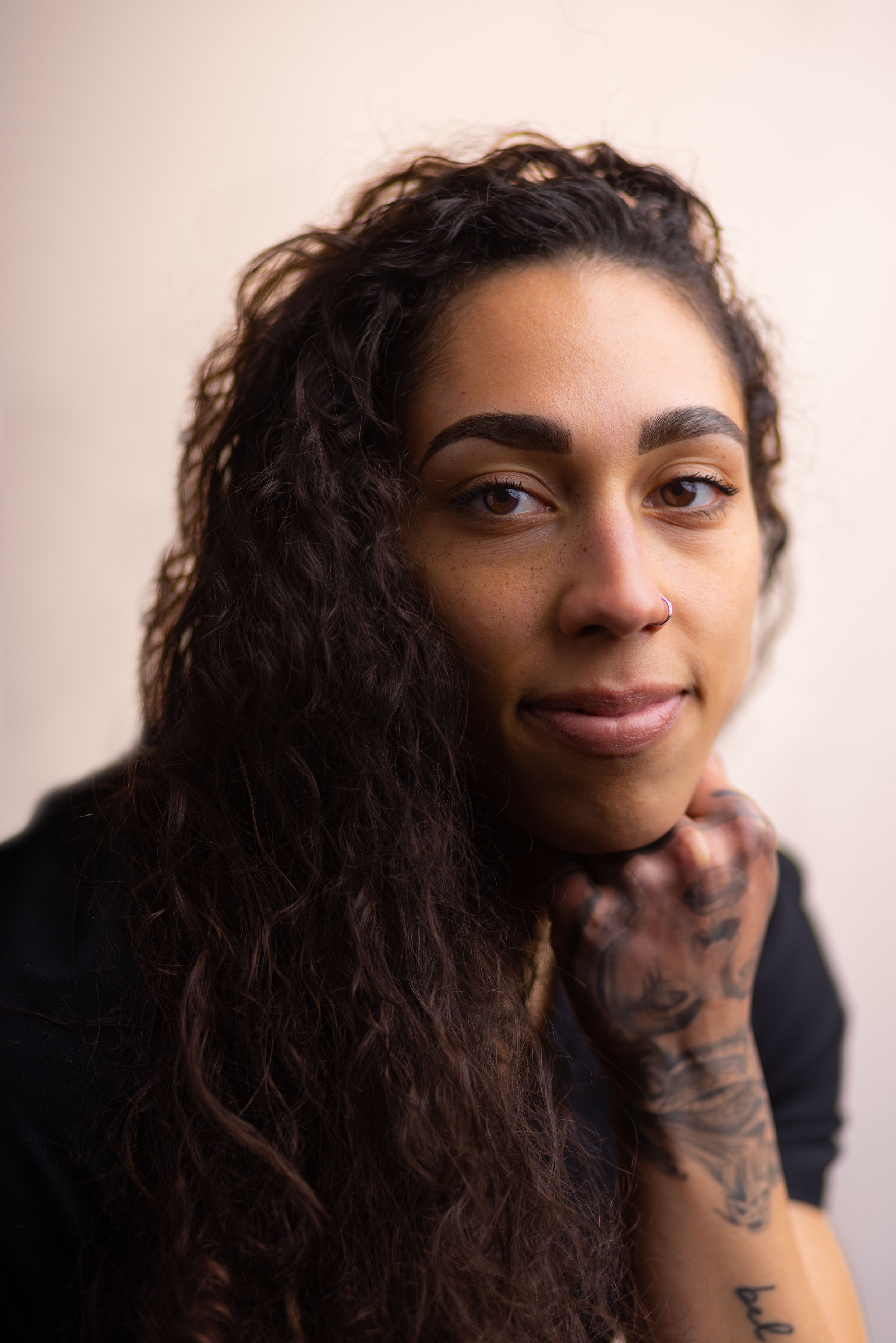 Close-up of a person with long curly hair, slightly smiling, hand tattoo visible on chin, neutral background.