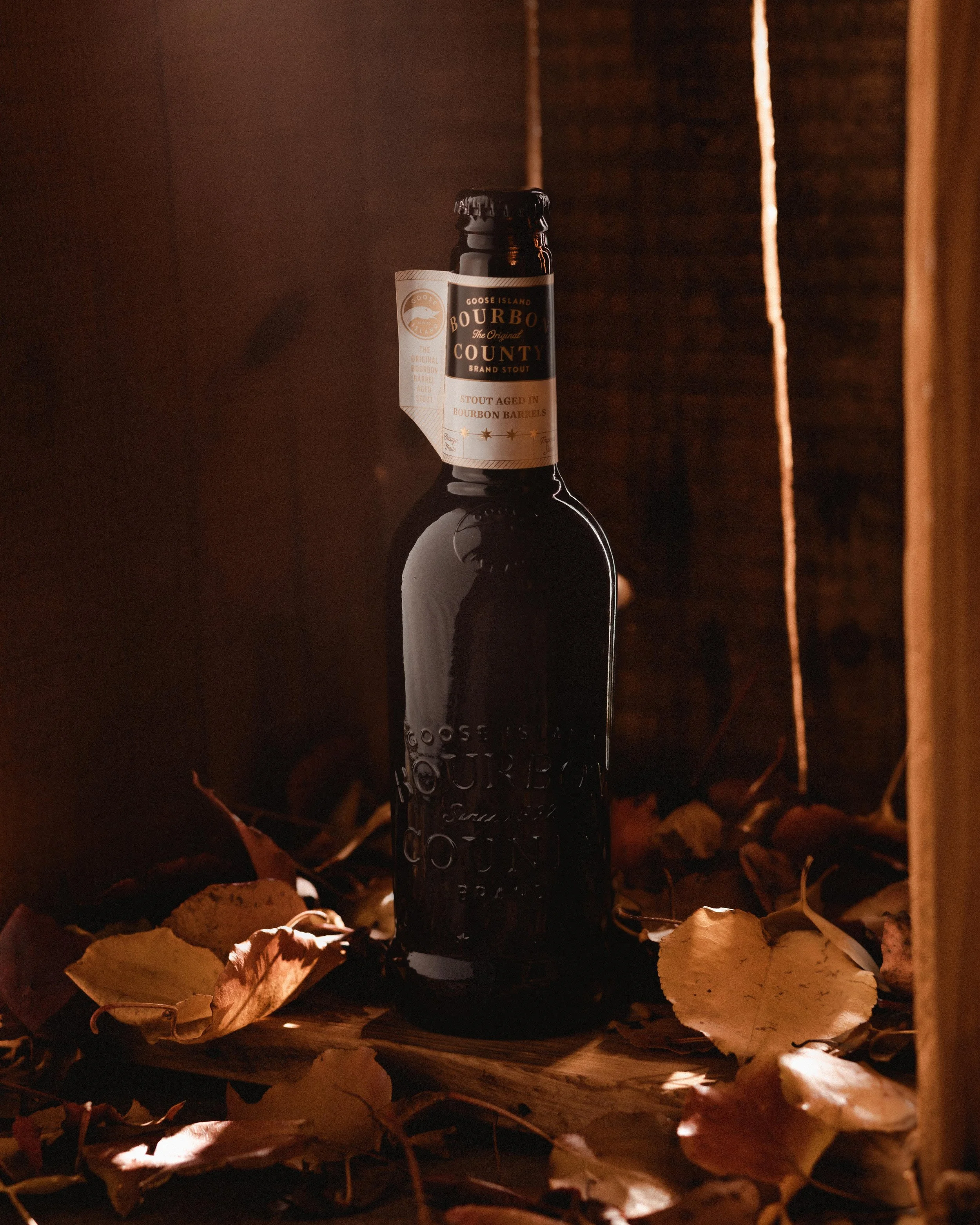 Bourbon County brand stout bottle surrounded by autumn leaves in a rustic wooden setting.