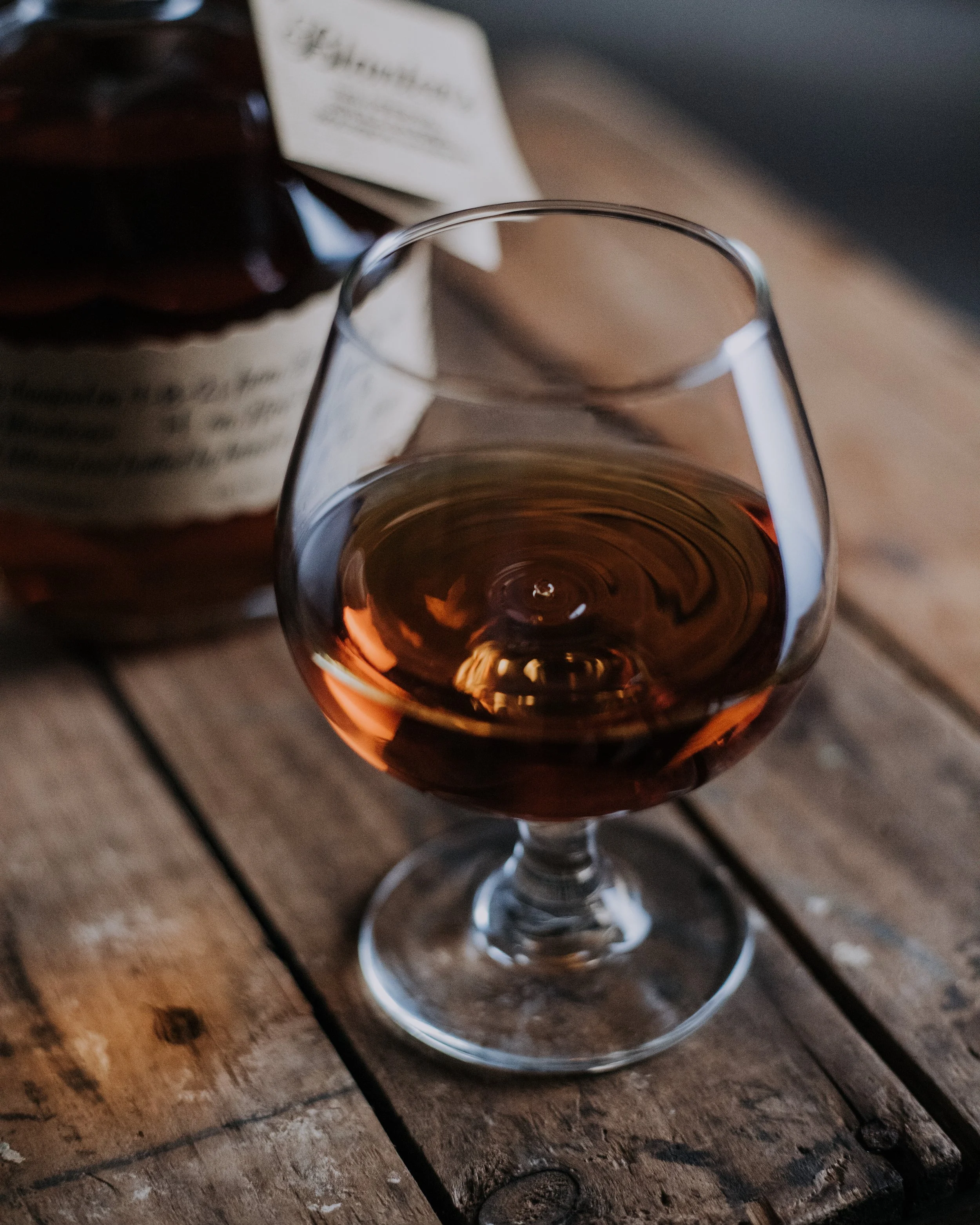A glass of amber-colored whiskey on a rustic wooden table, with a bottle in the background.