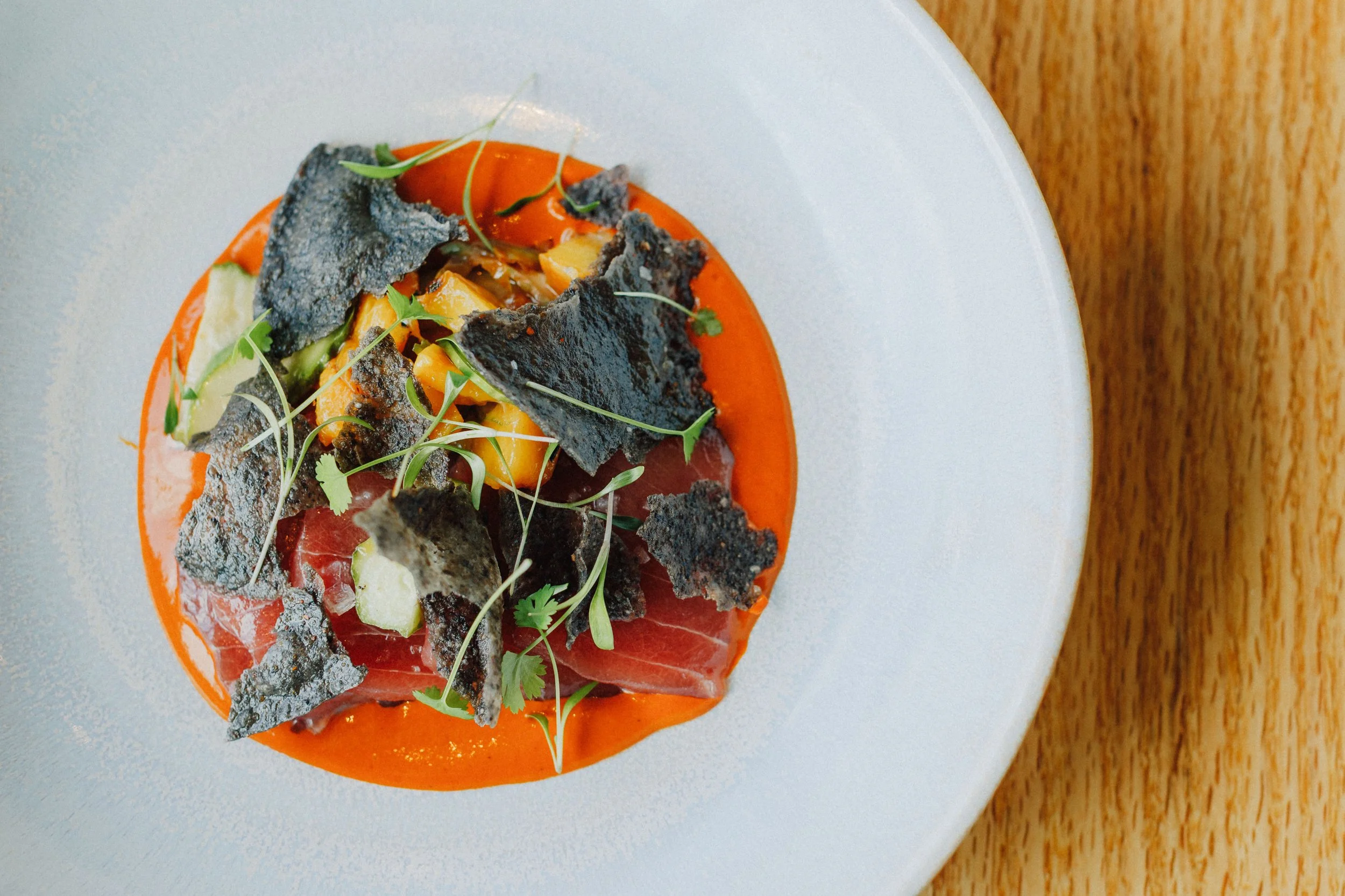 A gourmet dish with slices of raw beef, topped with black potato chips, microgreens, and thin slices of cucumber, served on a vibrant orange sauce on a white plate.