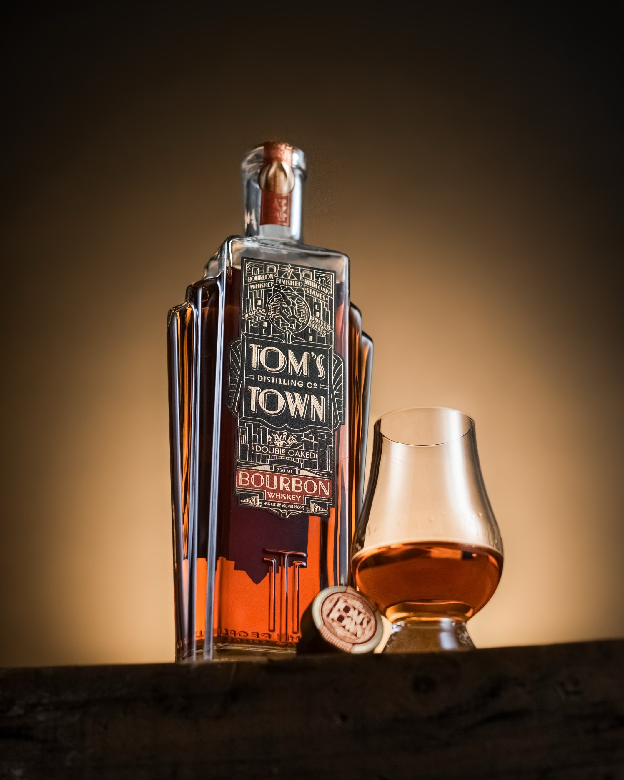 Bottle of Tom's Town bourbon whiskey with a glass and bottle cap on a wooden surface.