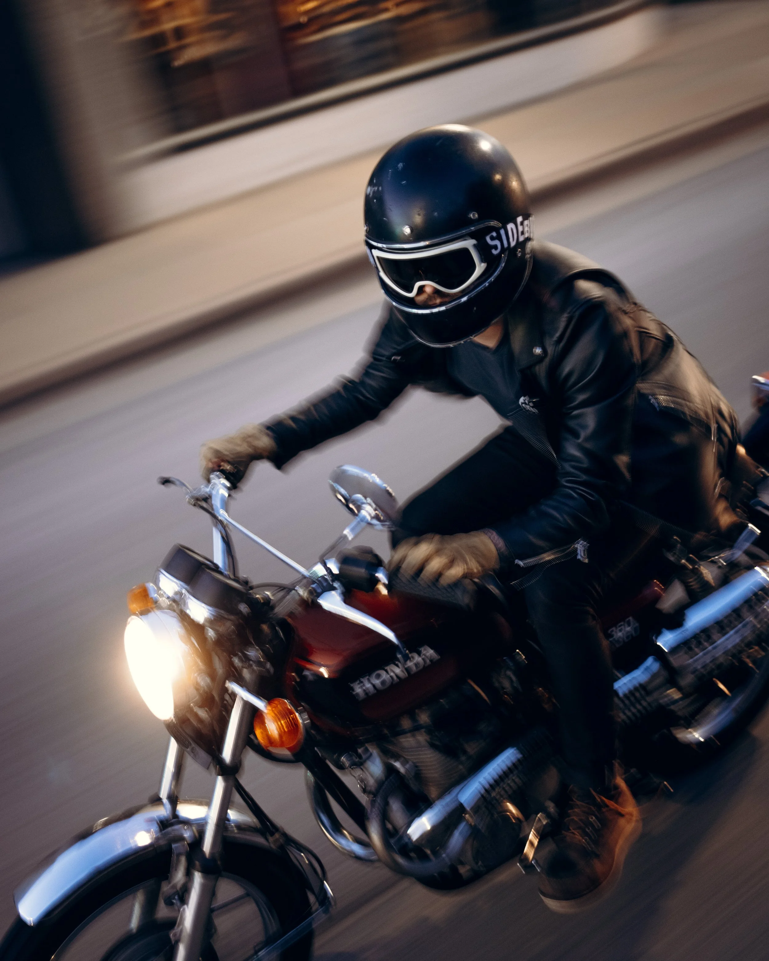 Motorcyclist wearing a black helmet and leather jacket riding a Honda motorcycle on the street.