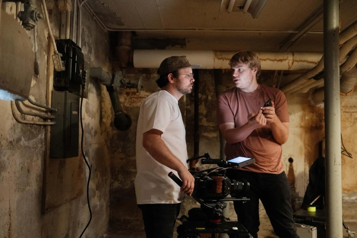 Two men with cameras and equipment are having a discussion in a basement with exposed pipes and pipes on the ceiling.