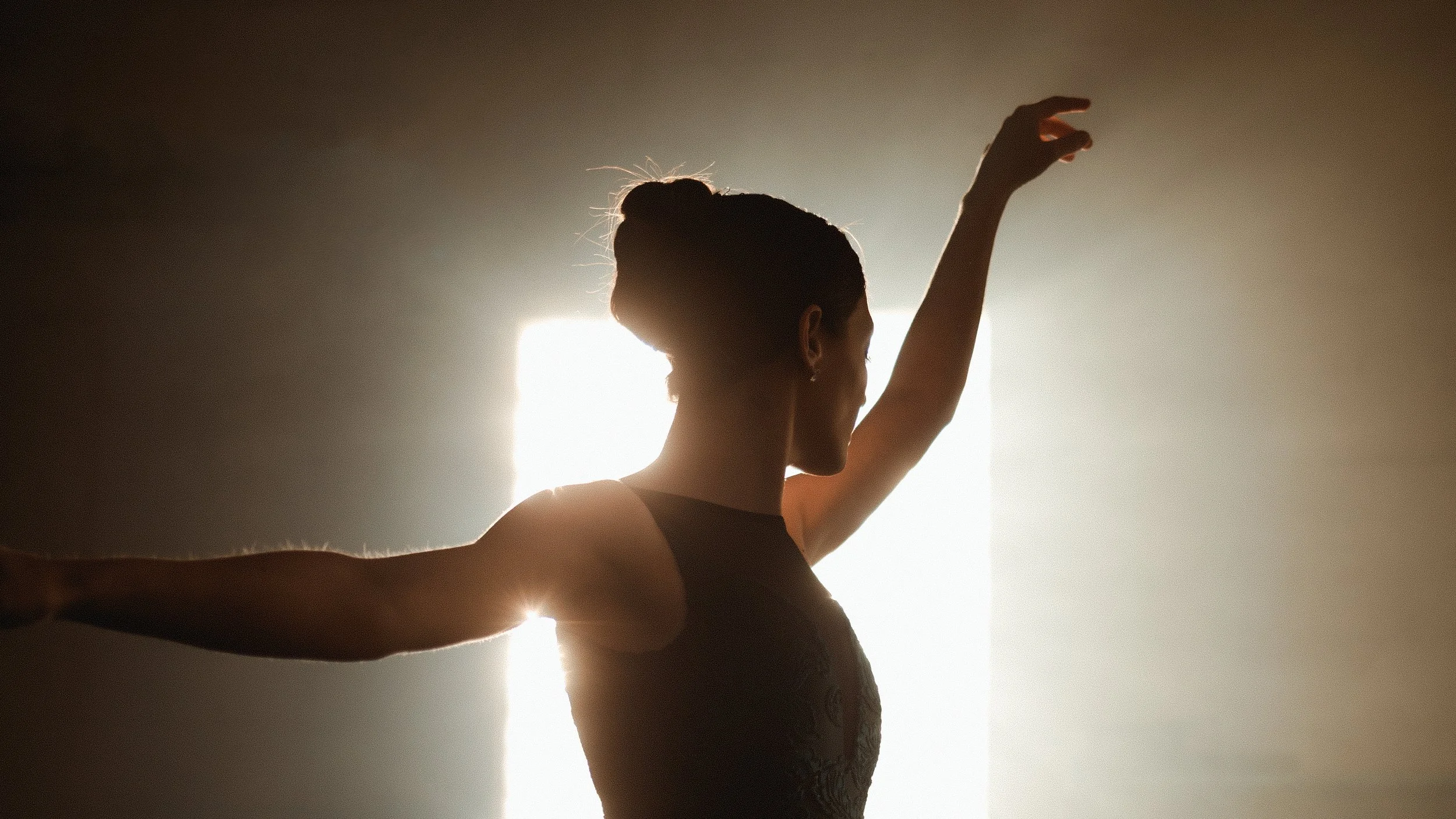 Silhouette of a ballet dancer with arms raised, backlit by bright light.