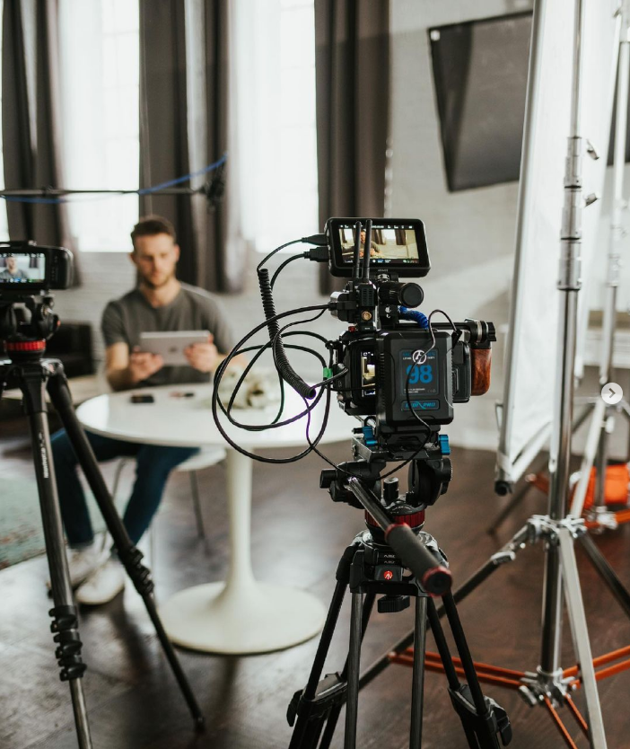 A man is sitting at a round table in a studio, using a tablet. There is professional video recording equipment, including cameras and lighting, set up around him for filming.