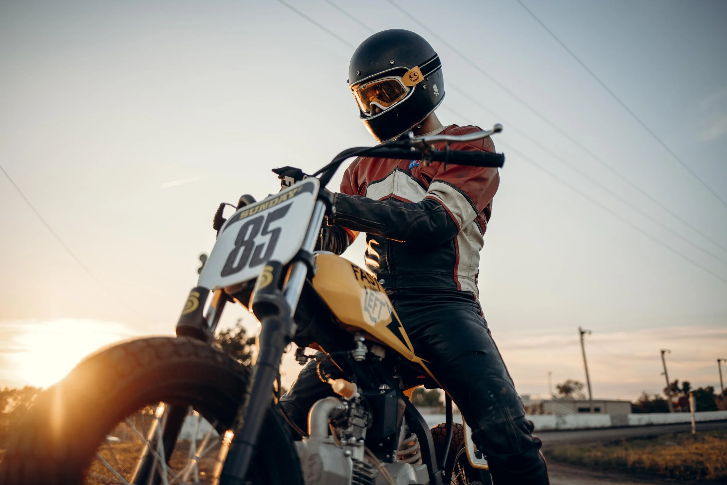 Motorcyclist wearing helmet and leather gear sitting on a dirt bike marked with the number 85, during sunset.