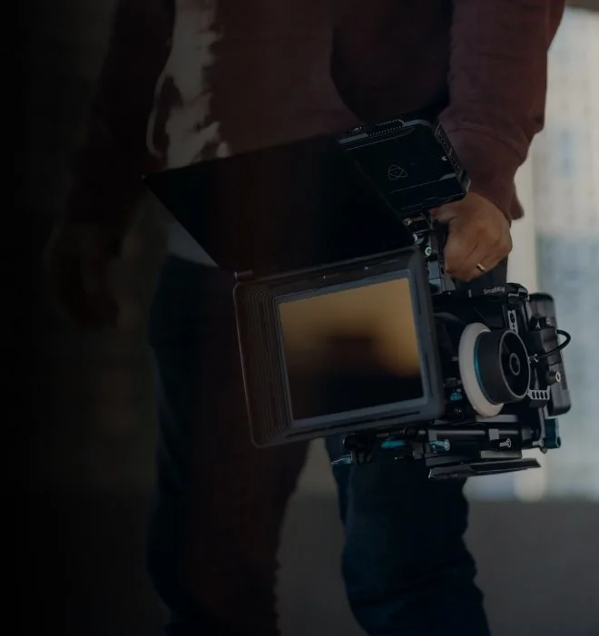 A person holding a professional camera with a large lens and a mounted monitor in a dimly lit room.