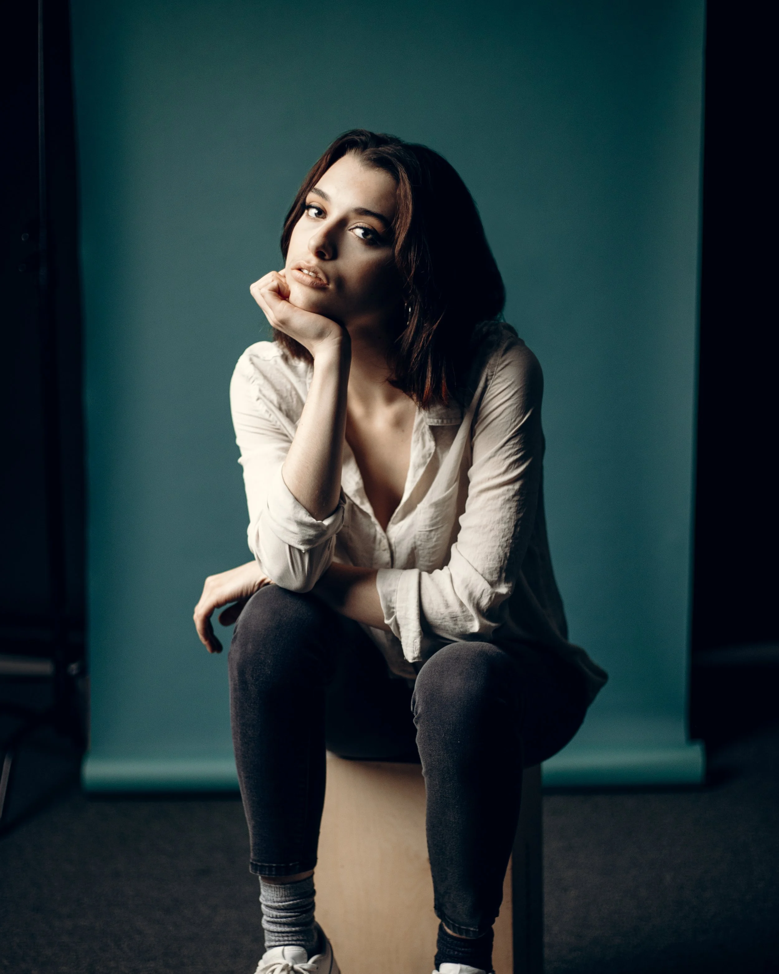 Person sitting on a stool, wearing a white shirt and jeans, posing thoughtfully with chin resting on one hand against a teal background.