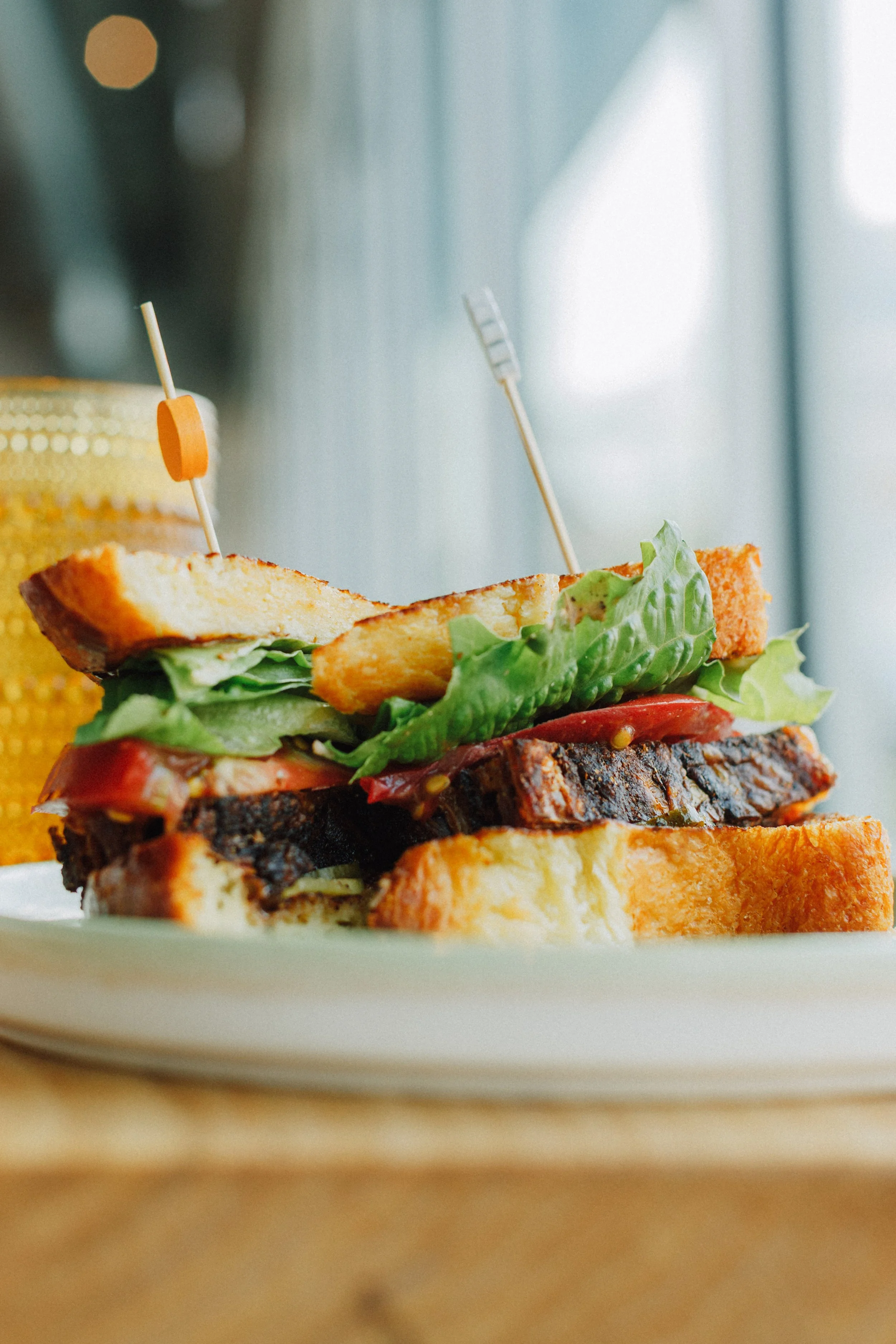 Close-up of a BBQ brisket sandwich with lettuce, tomato, toasted bread, and two toothpick skewers on a white plate.