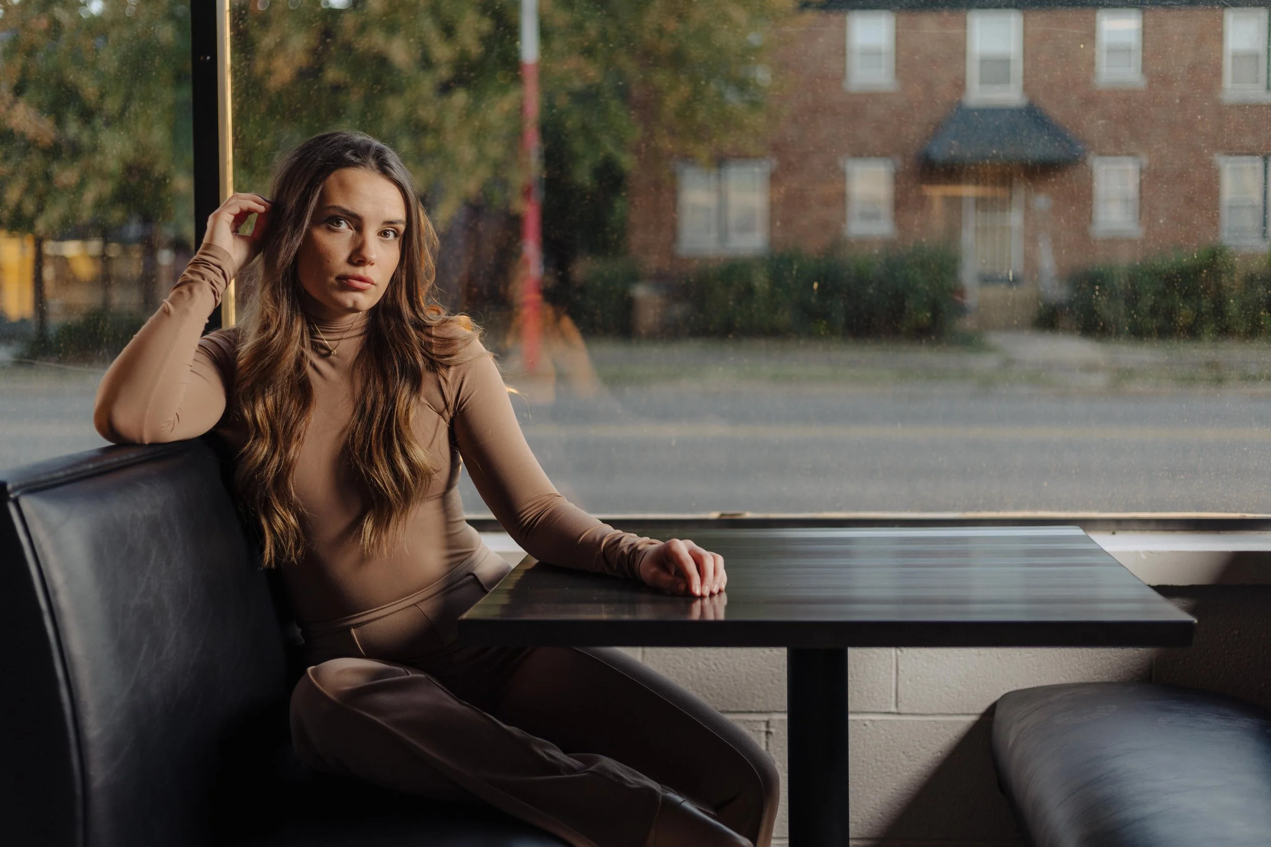 Woman sitting in a cafe booth by a window, wearing a taupe long-sleeve outfit, looking outside, with a contemplative expression.